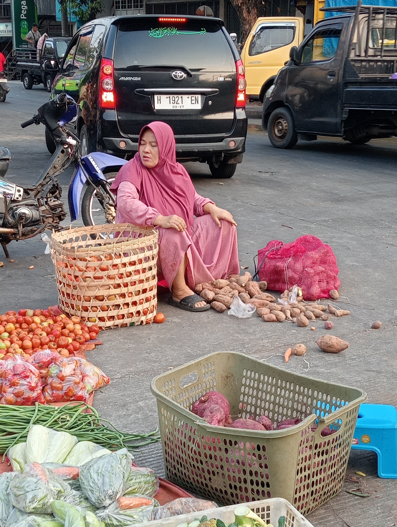The image depicts a street scene in Demak, Indonesia, likely a marketplace or roadside stall. A woman wearing a pink hijab and a matching pink dress is seated on the ground, next to a woven basket filled with tomatoes. She appears to be selling produce, as other vegetables like long beans, cabbage, and carrots are visible, some in plastic bags and some in baskets. A pile of yams or sweet potatoes is spread out on the ground in front of her, with more in a large red mesh bag and a green plastic basket. Several motorcycles are parked nearby, and various vehicles, including a black SUV, a yellow truck, and a black pickup truck, are present in the background, suggesting a busy street. The ground is paved, and the lighting indicates it is daytime, likely late afternoon given the shadows.