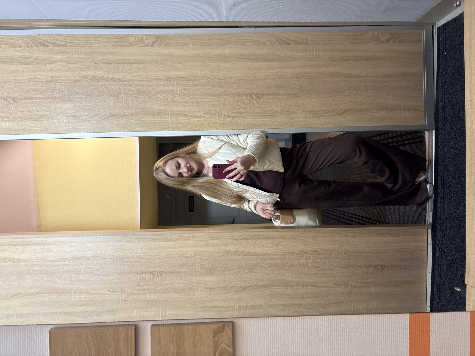 A young woman with long blonde hair and a pleasant, smiling expression is depicted taking a mirror selfie indoors in Chaikovsky, Russia. She is dressed in a light cream or off-white quilted jacket, a dark brown top, and matching dark brown wide-leg trousers. In her left hand, she holds a dark red or burgundy smartphone, while her right hand carries a beige handbag with a brown flap. A ring is visible on her left ring finger.
The mirror she is using forms part of a large wardrobe or sliding closet, distinguished by light wood-grain laminate panels framed in silver-colored metal. The reflection reveals the interior space behind her: a wall divided into two sections, one painted soft pink and the other bright yellow. Further in the background, a dark doorway is visible, above which a sign displays partially legible Cyrillic text, specifically "ПОМЕЩЕНИЕ" (meaning "ROOM" or "PREMISES"), likely part of a sign indicating a service or utility room.
The flooring in front of the mirrored wardrobe includes a dark grey or black textured carpet on the right side and a white and orange striped rug in the lower left portion of the image. Additionally, several light wood-like samples are arranged on the floor directly beneath the mirror. The ceiling overhead is white. The scene is well-lit, typical of an indoor environment, possibly a hallway or a room within a residence or hotel.