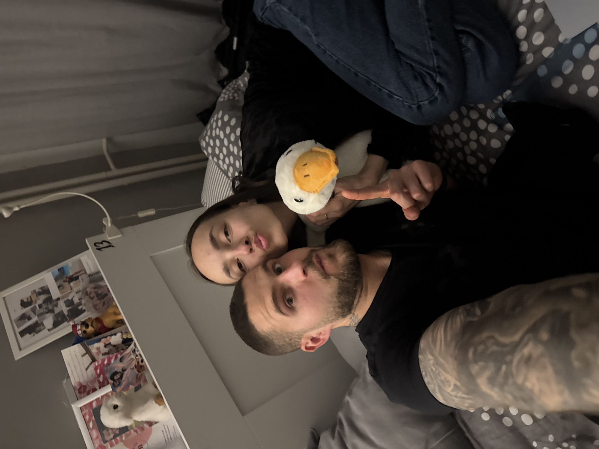 A medium shot shows a man and a woman lying in a bed with a plush goose toy, indoors. The man is on his side and wearing a black t-shirt. He has a short haircut and a beard. His arm has a tattoo. The woman is lying behind him with her head close to his. She is wearing a black top. Her hand is visible, pointing at the goose. The bed has a blue and white polka dot duvet cover. The location appears to be a bedroom, as seen by the photo frames. The number “13” is visible on a photo frame hanging on a wall. The location cannot be determined to be in the city of Sofia, Bulgaria.