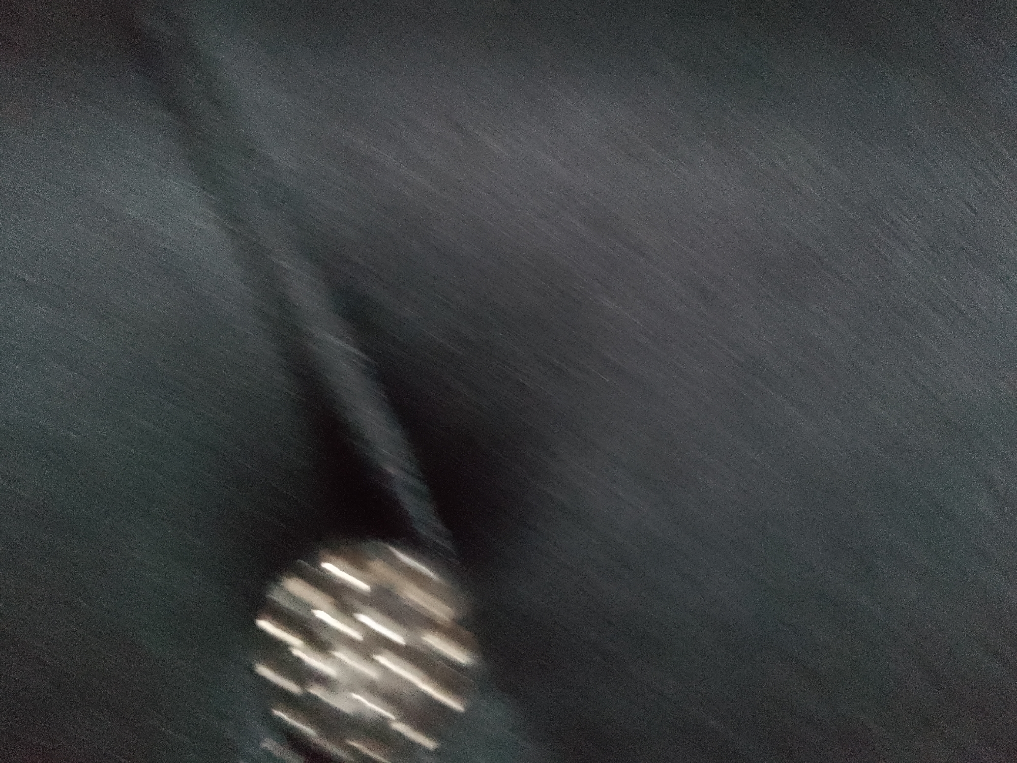 The image is an abstract, blurry shot of a dark surface with streaks of light. In the lower left quadrant, there is a blurred object that appears to be a metallic structure with multiple reflective facets, possibly a piece of machinery or jewelry. The surface itself has a fine texture and appears to be made of dark material, possibly fabric or a rough painted surface. The lighting suggests a low-light environment with a moving light source, creating the motion blur effect. There is no discernible location in Jauro Yinu, Nigeria, or any specific environmental context within the image.
