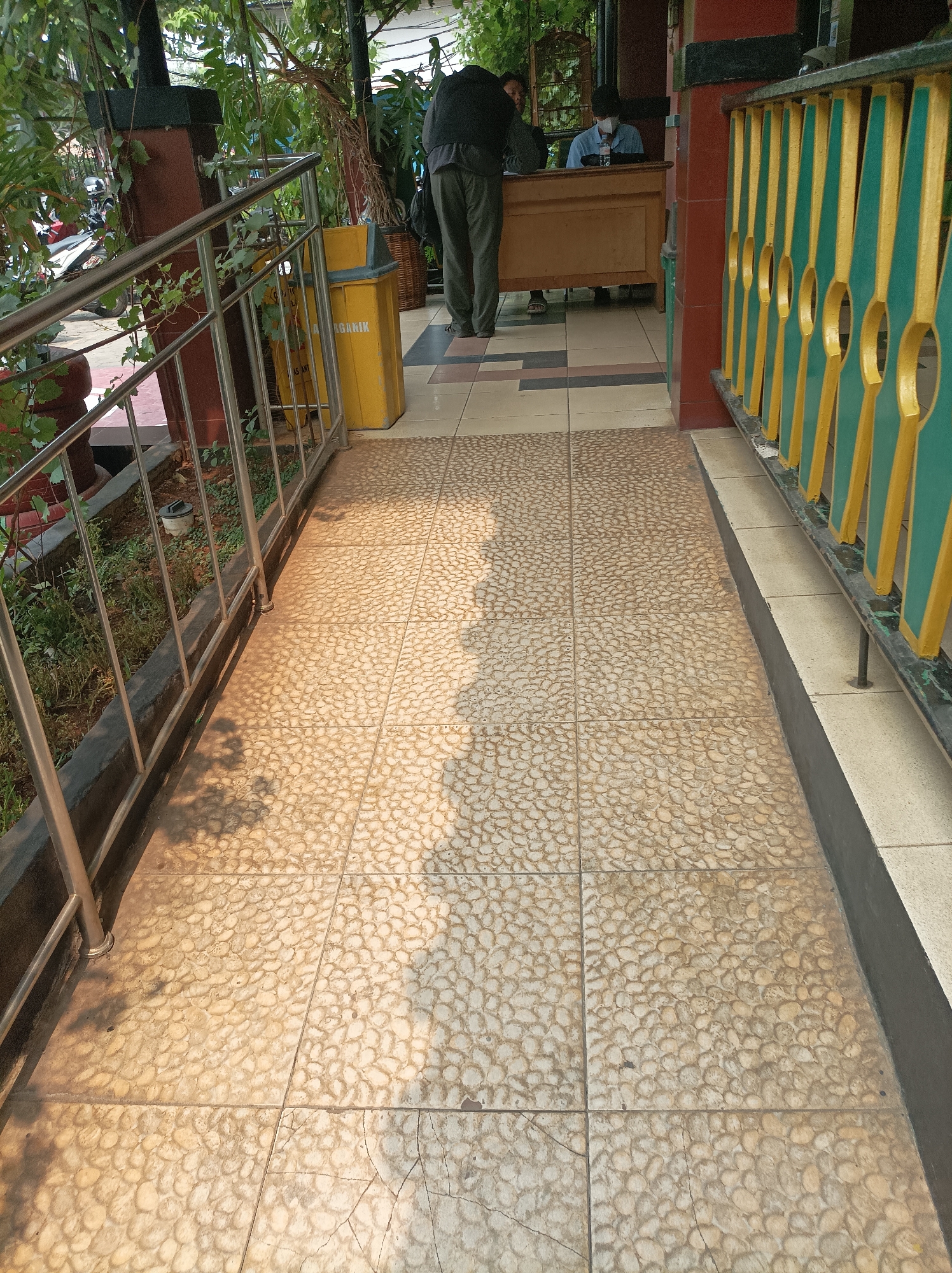 The image depicts an outdoor walkway in West Jakarta, Indonesia, likely in front of a building with an office or service area. The walkway is paved with small, light-colored pebbles embedded in concrete, creating a textured surface. Sunlight casts shadows from the foliage and a metal railing onto the walkway, indicating it is daytime.
To the left, a metal railing runs along a raised planter bed containing greenery and soil. A yellow trash bin with "ORGANIK" written on it is visible next to the railing. Further back, a person wearing dark clothing stands with their back to the camera, near a woven basket.
To the right, a colorful railing with alternating green and gold vertical elements separates the walkway from a raised area. In the background, two people are visible. One, wearing a light blue shirt and a face mask, sits behind a wooden counter, appearing to be an employee. Another person, partially obscured, stands in front of the counter, possibly interacting with the employee. The overall environment suggests a public space, possibly a government office or a community center.