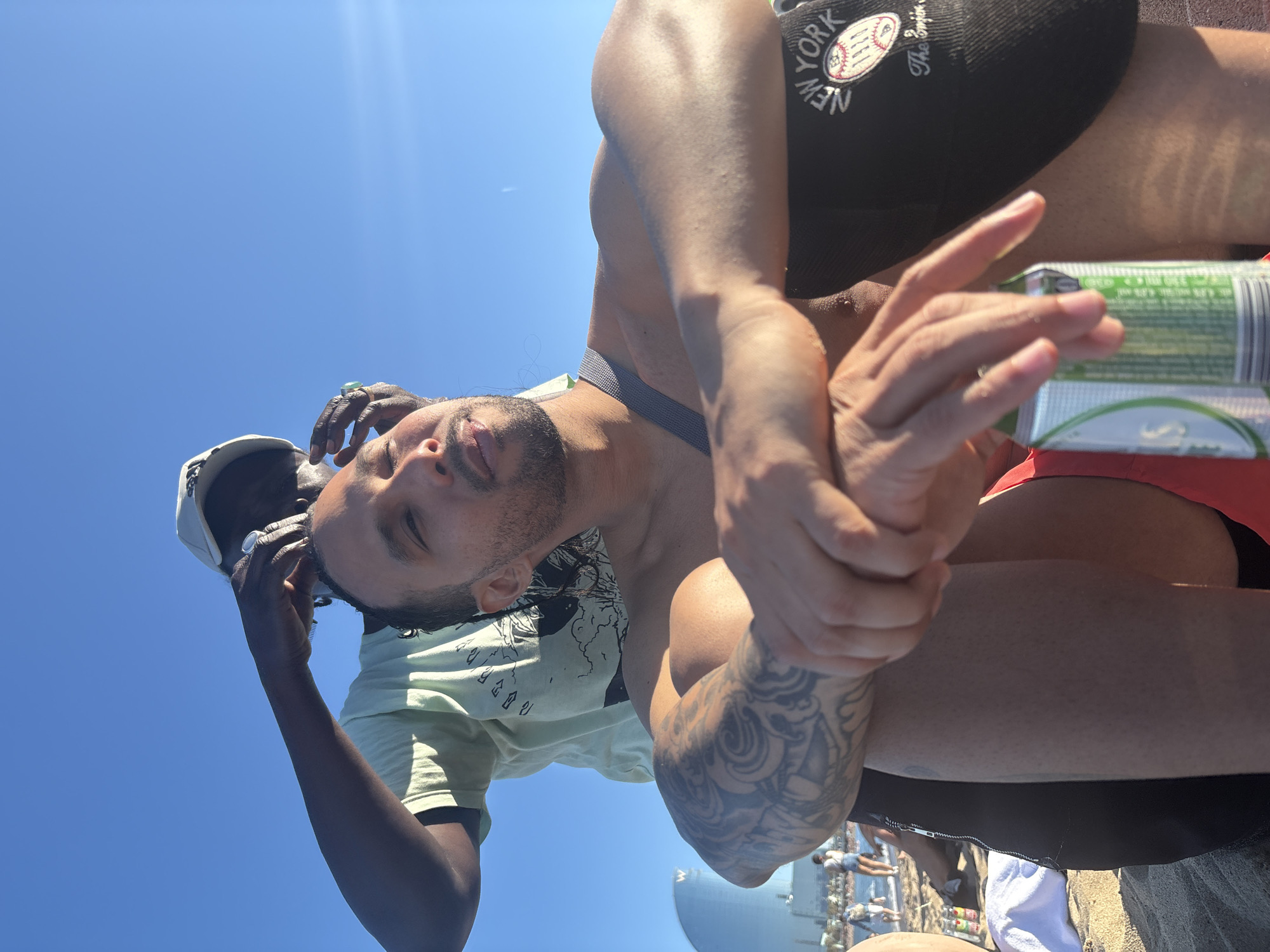 A low-angle shot shows two men on a sunny day. One man, lighter-skinned with a beard, is wearing a black New York baseball cap, a grey harness, and has a tattoo on his arm. He holds a pack of cigarettes. The other man, darker-skinned, wears a light green t-shirt and a white baseball cap and has a ring on his finger. In the background, there is a group of people, a large metal tank, and the city of Barcelona can be assumed to be the location due to its recognizable structure in the backdrop. The sky is clear blue.