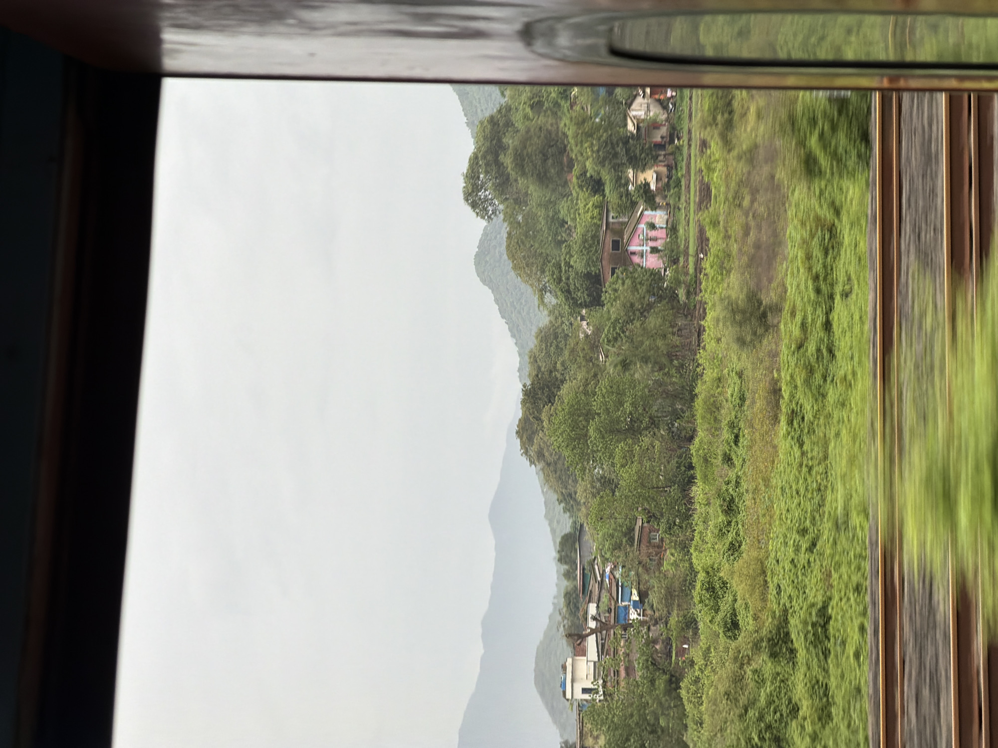 The image captures a view from a moving train window, showing a village nestled amidst lush green hills under a cloudy sky. In the foreground, blurred train tracks run horizontally across the frame. Beyond the tracks, dense, vibrant green foliage covers the landscape, with a small village scattered on the slopes of the hills. The village consists of several houses, some with tiled roofs and brightly colored facades, including a distinctive pink house. In the background, layers of hazy, green mountains recede into the distance. The overall scene suggests a rural or semi-rural setting, likely in the outskirts of Vasai-Virar, India, characterized by its natural beauty and modest human settlements. The overcast sky indicates a potentially damp or humid climate.