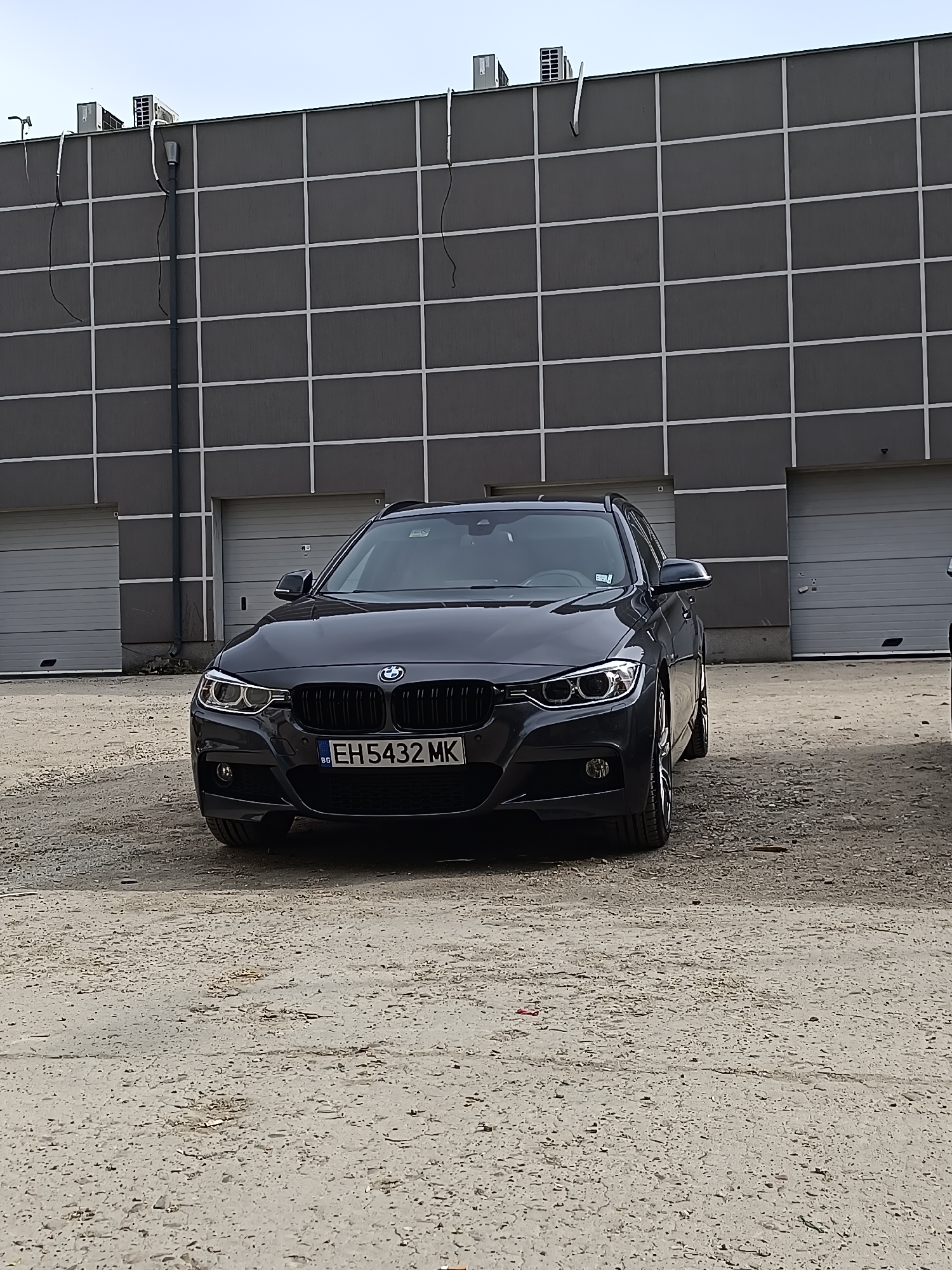 The image shows a dark grey BMW 3 Series Touring station wagon parked on a rough, concrete surface, likely a parking lot or industrial area. Behind the car is a large, grey, industrial building with a grid-like pattern and several closed garage doors. On the top of the building there are multiple air conditioning units. The car's license plate reads "EH5432 MK," indicating it is registered in Bulgaria. There are no people visible in the image. Based on the license plate, the location is presumed to be in Bulgaria, though the specific city (Pleven) cannot be directly inferred from the image.