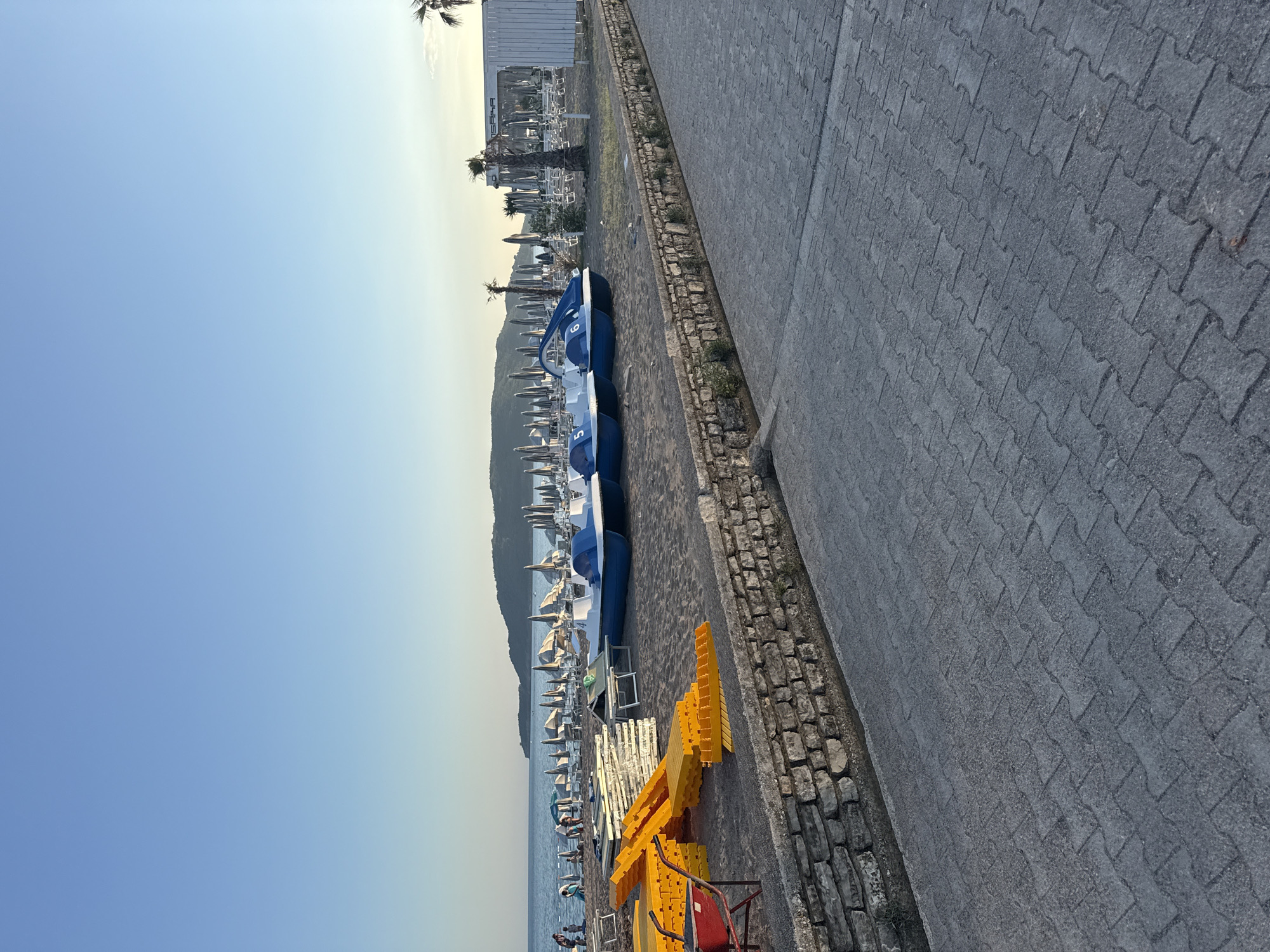 The image depicts a beach scene in Bečići, Montenegro, likely during the late afternoon or early evening, given the soft light. The foreground features a paved walkway constructed of interlocking grey pavers, bordered by a low stone wall. Beyond the wall lies a sandy beach area. On the sand, several blue and white paddle boats are parked. To the left, a stack of yellow plastic platform pieces and a red wheelbarrow are present, suggesting preparation or disassembling of beach infrastructure. In the background, numerous beach umbrellas are set up, with some lounge chairs visible beneath them. The sea is calm and extends to the horizon, where a hilly coastline is visible. A white building with a sign that appears to read "SASA" is situated towards the right side of the image, with palm trees planted in front of it. Several people are visible on the beach in the middle ground, some walking and others relaxing. The sky is a clear, pale blue, with a gradient towards a lighter hue near the horizon.
