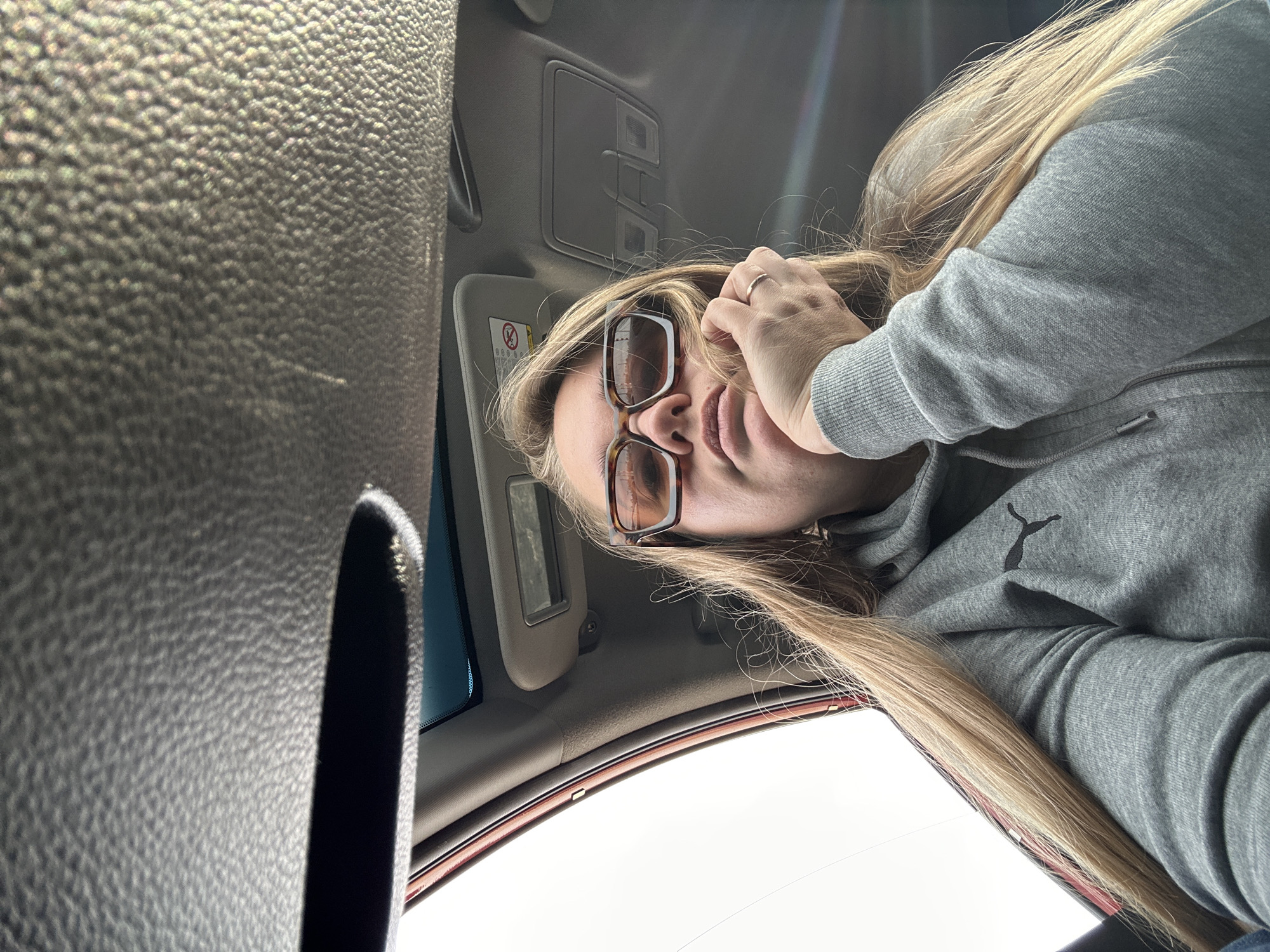 A photograph depicts a person inside a vehicle, likely a car, with the camera positioned from below. The person, a woman, is wearing glasses and a gray hooded sweatshirt, and has long blonde hair. She is holding her hand near her face. The interior of the vehicle includes a sun visor, a rearview mirror, and a textured dashboard. The background shows the outside environment visible through the car windows, appearing bright. The image provides no specific information to identify the location in Ural'skoe, Russia.