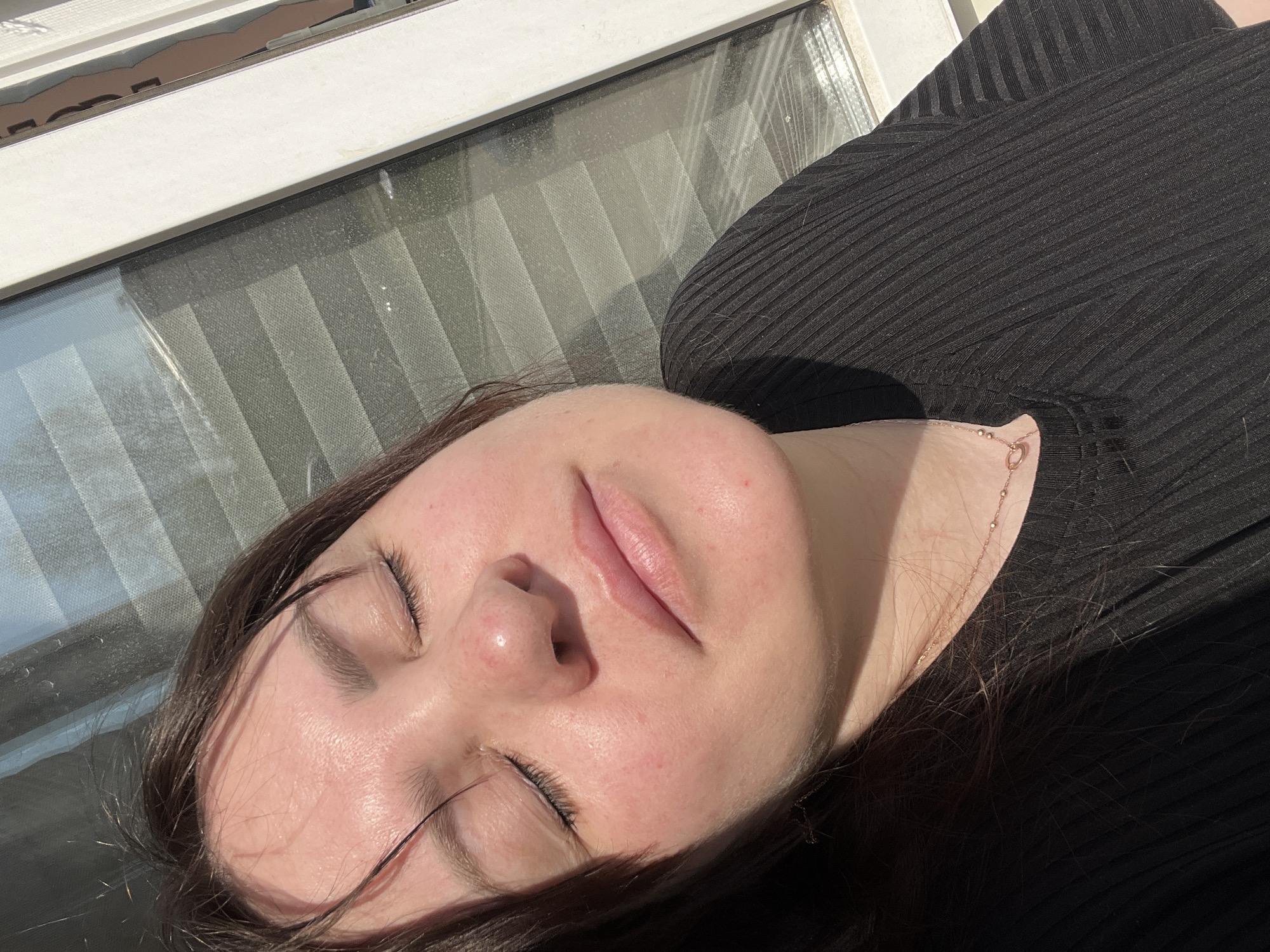A young woman with closed eyes lies in the sunlight, her face and upper chest visible. She has fair skin with some redness on her cheeks and nose. Her dark hair is visible around her face and neck. She wears a black, ribbed top, and a delicate gold necklace adorns her neck. Behind her is a window with blinds. The image does not provide sufficient information to locate the scene in the city of Noviy, Russia.