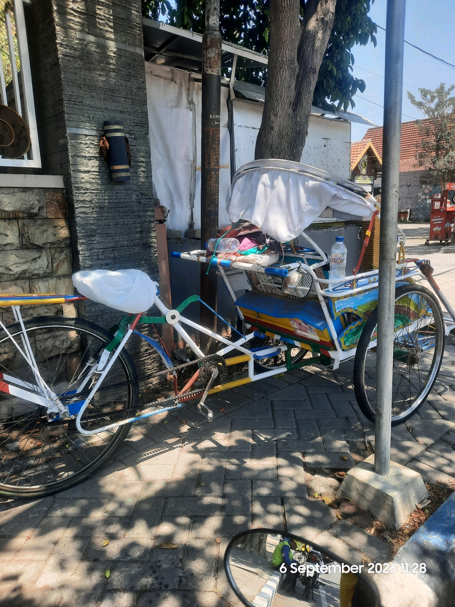 The image depicts a brightly colored pedicab parked on a cobblestone sidewalk in Probolinggo, Indonesia. The pedicab, which is white with colorful accents of red, yellow, blue, and green, has a covered passenger seat and a small engine. A plastic bottle of water is visible on the passenger seat. A tall, weathered pole stands to the right of the pedicab. In the background, there are buildings with red tile roofs and trees with green leaves. The scene is illuminated by sunlight, casting shadows on the ground. The date and time stamp "6 September 2026 11.28" is visible in the bottom right corner, overlaying a reflection in a mirror.