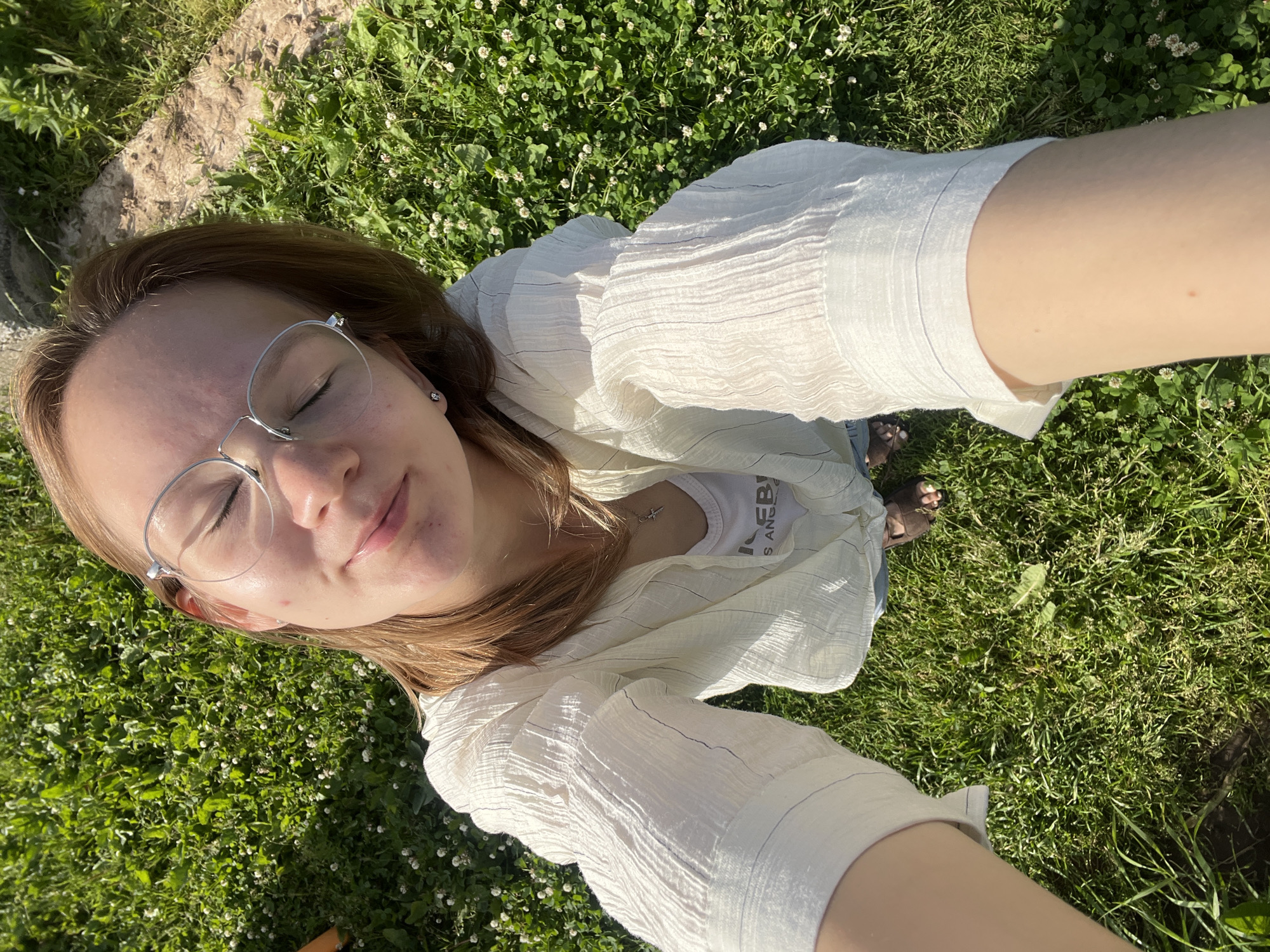 A young woman with blonde hair, wearing glasses, a white linen shirt over a white tank top, and blue shorts, is lying on a grassy field. The sun is shining, casting shadows on the grass, suggesting it is daytime. The field is covered in lush green grass and small white clover flowers. The woman's eyes are closed, and she appears to be relaxed and enjoying the outdoors. Her right hand is outstretched, holding the camera, indicating she is taking a selfie. The background is blurred, but hints of a stone structure and more grass suggest a natural, outdoor environment. A faint inscription is visible on her tank top, though it is partially obscured. The overall impression is one of peacefulness and enjoyment of a sunny day in a natural setting.