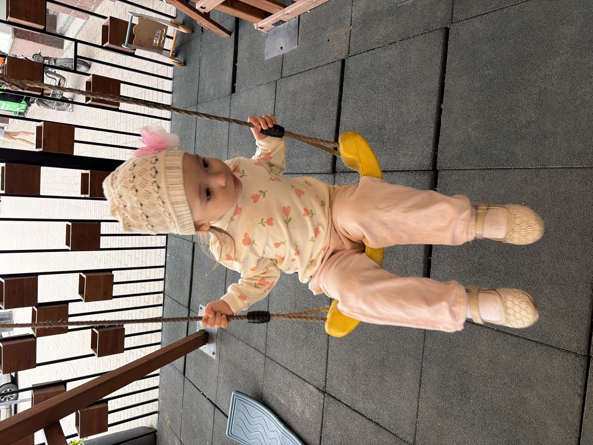 A young child is swinging on a playground. The child, wearing a beige knit hat with a pink flower, a long-sleeved shirt with a heart pattern, pink pants, and light-colored shoes, is holding onto the swing ropes. The swing seat is yellow. The setting appears to be an outdoor playground with a rubberized safety surface. In the background, there are wooden structures and a fence with a white slatted design. Visible in the distance is a bicycle and a green cart. The lighting suggests it is daytime, with overcast weather. The child's gaze is directed downwards, possibly indicating enjoyment or concentration. No visible text is present in the image.