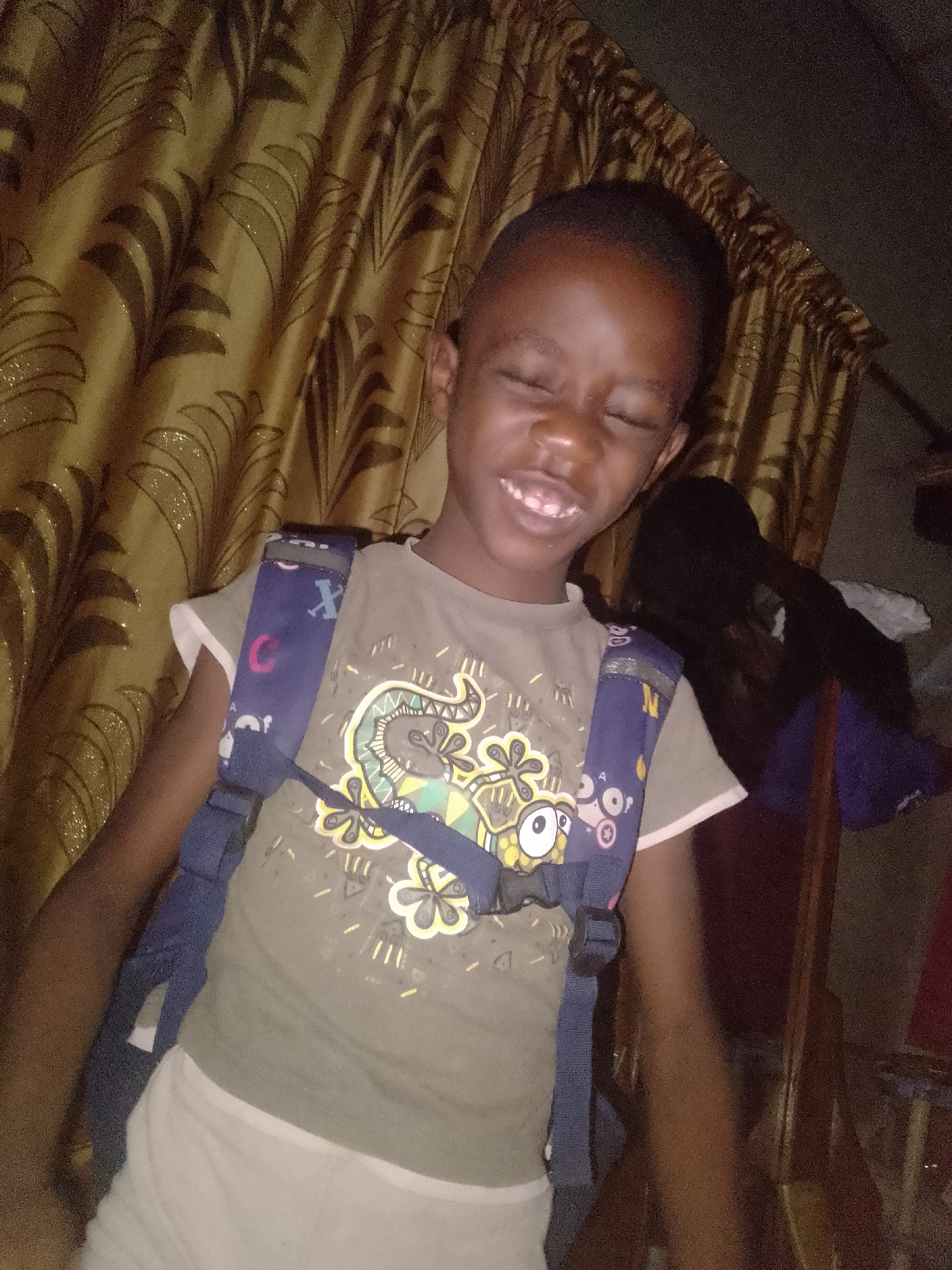 A young Black child is seen indoors, wearing a backpack and a shirt with a lizard-like creature graphic. They are smiling with their eyes closed. Behind them is a curtain with a patterned design and other indiscernible objects. The location appears to be Unknown, Unknown.