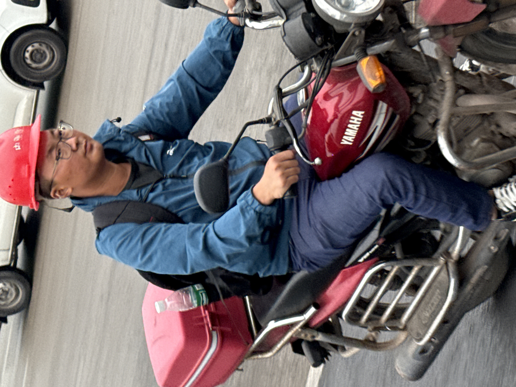 A man wearing a red cap and glasses is riding a Yamaha motorcycle on a road, likely in Jiangjin, China. He is wearing a blue jacket and pants, and has a backpack on. The motorcycle is red and black, and there's a red cargo box on the back carrier. The setting appears to be an urban street. A white vehicle can be partially seen on the left side of the image.