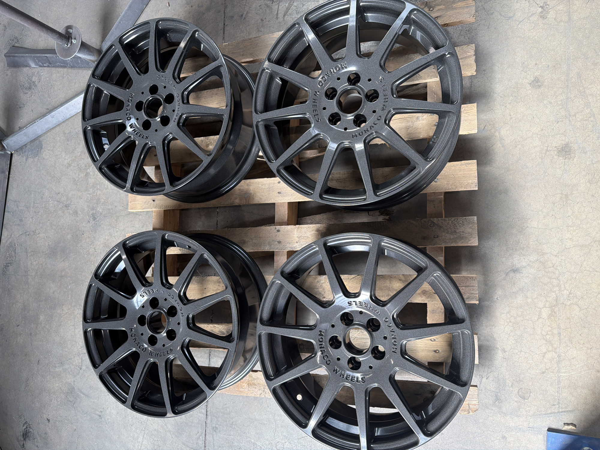 Four dark gray, multi-spoke alloy wheels are arranged on a wooden pallet on a concrete floor. The wheels appear to be new, with a metallic finish. Each wheel has "MONACO WHEELS" embossed around the center hub. The background consists of a concrete floor and some industrial metal structures. The setting is likely a workshop or storage area.