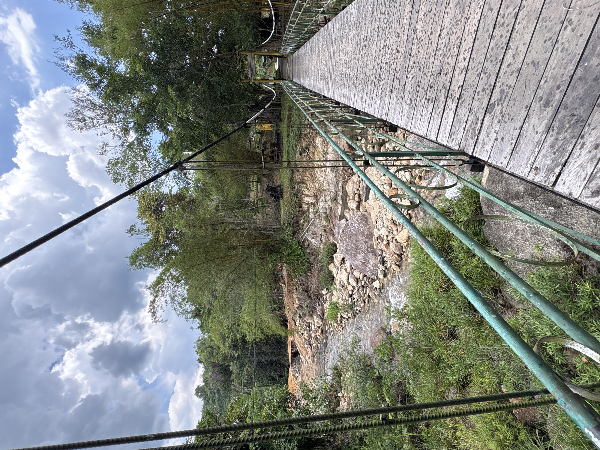 A low-angle, close-up captures the perspective of someone standing on a wooden suspension bridge in Khanh Vinh, Vietnam. The bridge has green metal railings with decorative circular elements. Below, a rocky stream bed contains a narrow stream of water. Lush green vegetation lines the banks and extends into a dense forest in the background. Overhead, a partly cloudy sky is visible. Thick cables support the bridge.