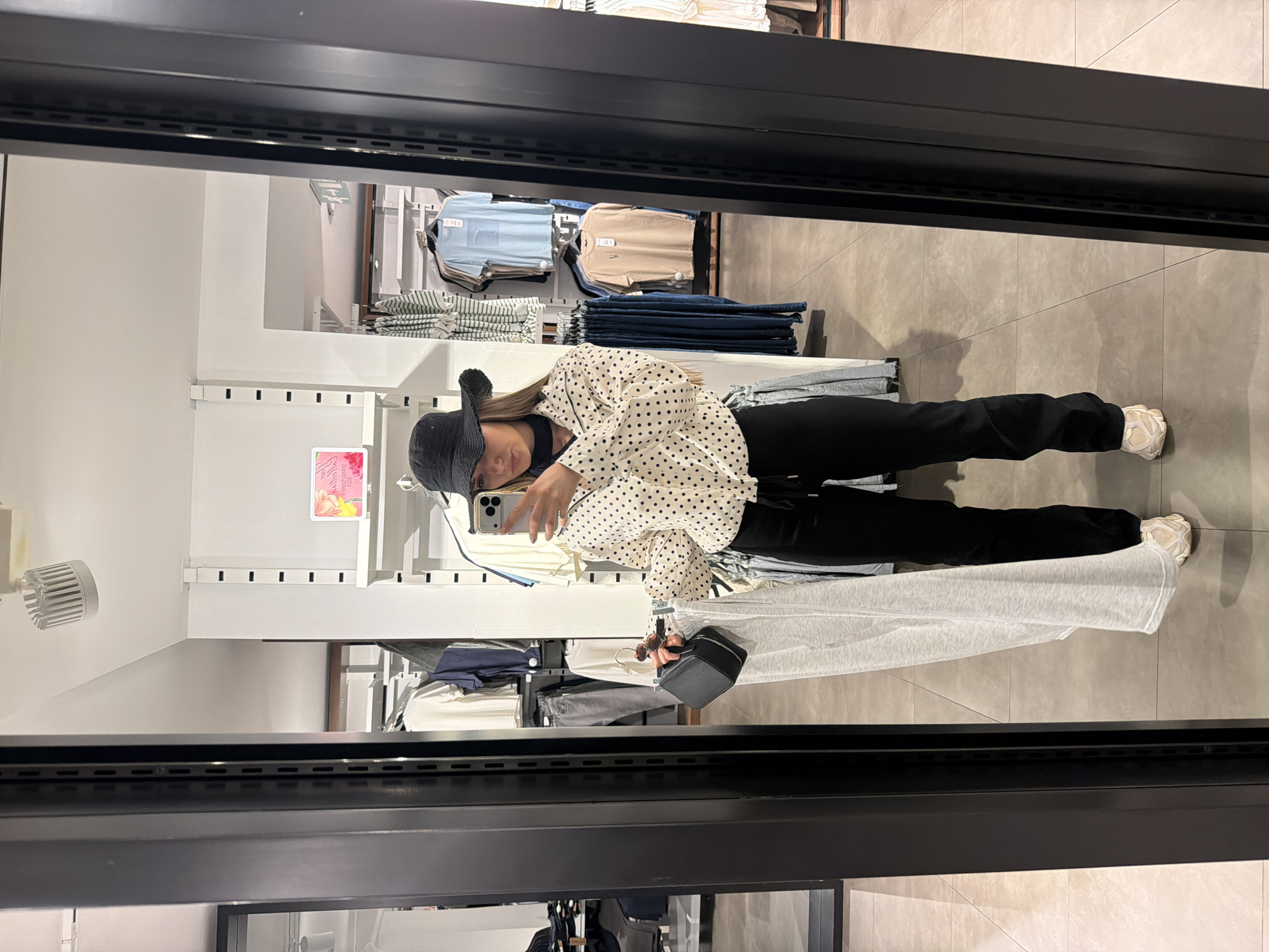 A woman in a black hat and polka dot blouse is taking a selfie in front of a mirror in what appears to be a clothing store. She is holding her phone in her left hand and a small black purse in her right. She is wearing black pants and white sneakers. Clothes racks are visible in the background with various items of clothing displayed on them. The floor is made of light grey tiles. Based on the clothing on the racks and the overall appearance, the location could be a shop or department store in the city of Sofia, Bulgaria.