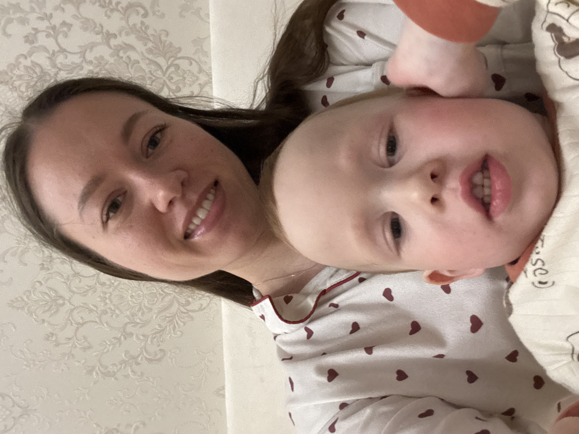 A woman with long brown hair is smiling and looking at the camera while lying next to a toddler. The toddler is on their side, also looking at the camera with a smile. Both are wearing pajamas with red heart patterns. The background is a light-colored wall with a floral pattern. The location is potentially a domestic setting in Ферма, Russia.