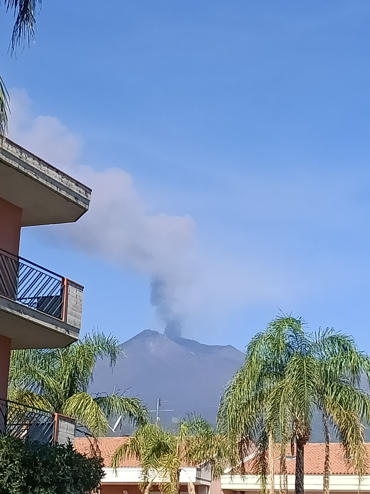 A volcano is erupting in the background, emitting a plume of smoke that rises into the clear blue sky. In the foreground, a multi-story building with a balcony and a red facade is visible. Palm trees are scattered throughout the scene, with their fronds reaching towards the sky. The building appears to be residential or commercial, with tiled roofs and visible balconies. The overall atmosphere is one of natural power and potential danger, juxtaposed with the everyday scene of a building and vegetation.