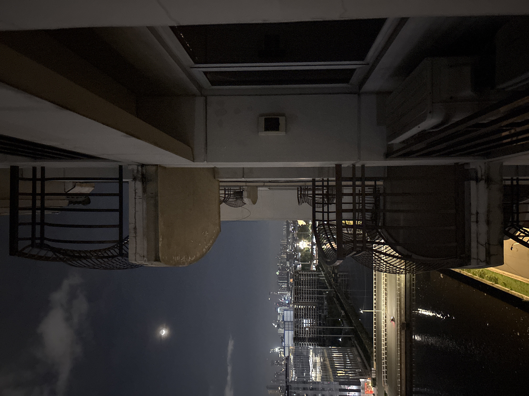 The image is a night-time view from a high-rise building in Pasay, Philippines. A sliver of the crescent moon is visible in the dark sky, with some clouds. Below, a cityscape unfolds with numerous buildings, some illuminated by lights, suggesting a densely populated urban area. A body of water, likely a river or bay, reflects the city lights. A road runs alongside the water, with a single vehicle visible on it, its headlights illuminating the path. The foreground shows a balcony railing, partially obscuring the view, and a portion of the building's exterior, including a window and an air conditioning unit. The overall impression is of a dynamic, nocturnal urban environment.