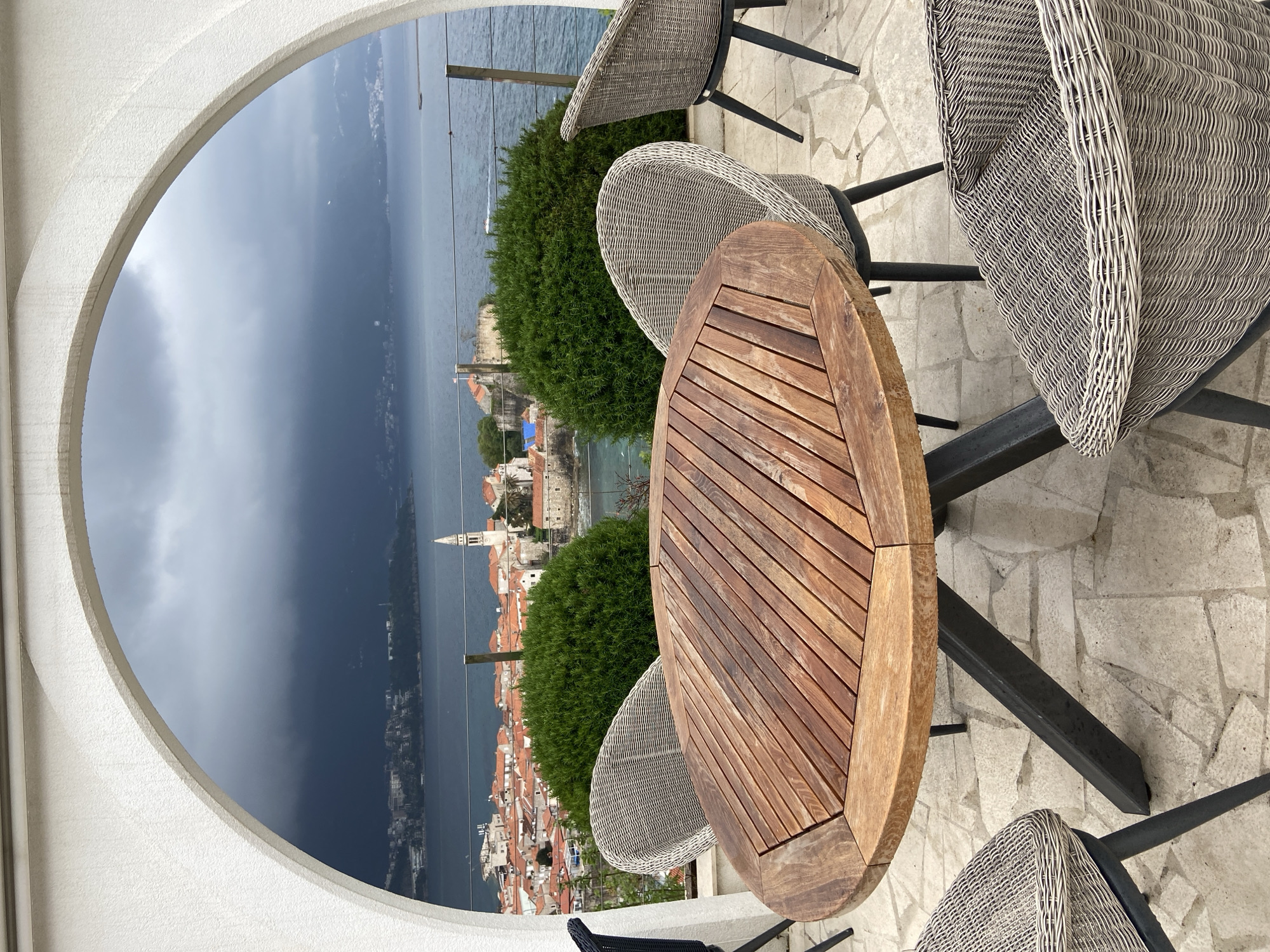 Here is a detailed description of the image:
**General Scene:**
The image shows an outdoor terrace or patio setting, with a view overlooking the city of Budva, Montenegro, from an elevated position. The weather appears to be overcast, possibly with a storm approaching.
**Objects:**
* **Table:** A wooden, oval-shaped table is centered in the foreground. It has a slatted surface.
* **Chairs:** Wicker or rattan-style chairs surround the table.
* **Bushes:** Two rounded, green bushes are strategically placed to enhance the outdoor setting.
* **Arch:** A white, rounded arch frames the view of Budva and the sea.
* **Cityscape:** Budva is visible in the distance, showcasing its characteristic red-tiled roofs and the tower.
* **Sea/Bay:** A body of water, likely part of the Adriatic Sea, stretches out in the background.
* **Sky:** The sky is filled with dark, ominous clouds, suggesting an approaching storm.
**Location:**
The viewpoint is from an elevated terrace or balcony overlooking Budva, Montenegro.
**Environment:**
The terrace has a stone-paved surface. The overall environment suggests a relaxing or dining area with a scenic view.