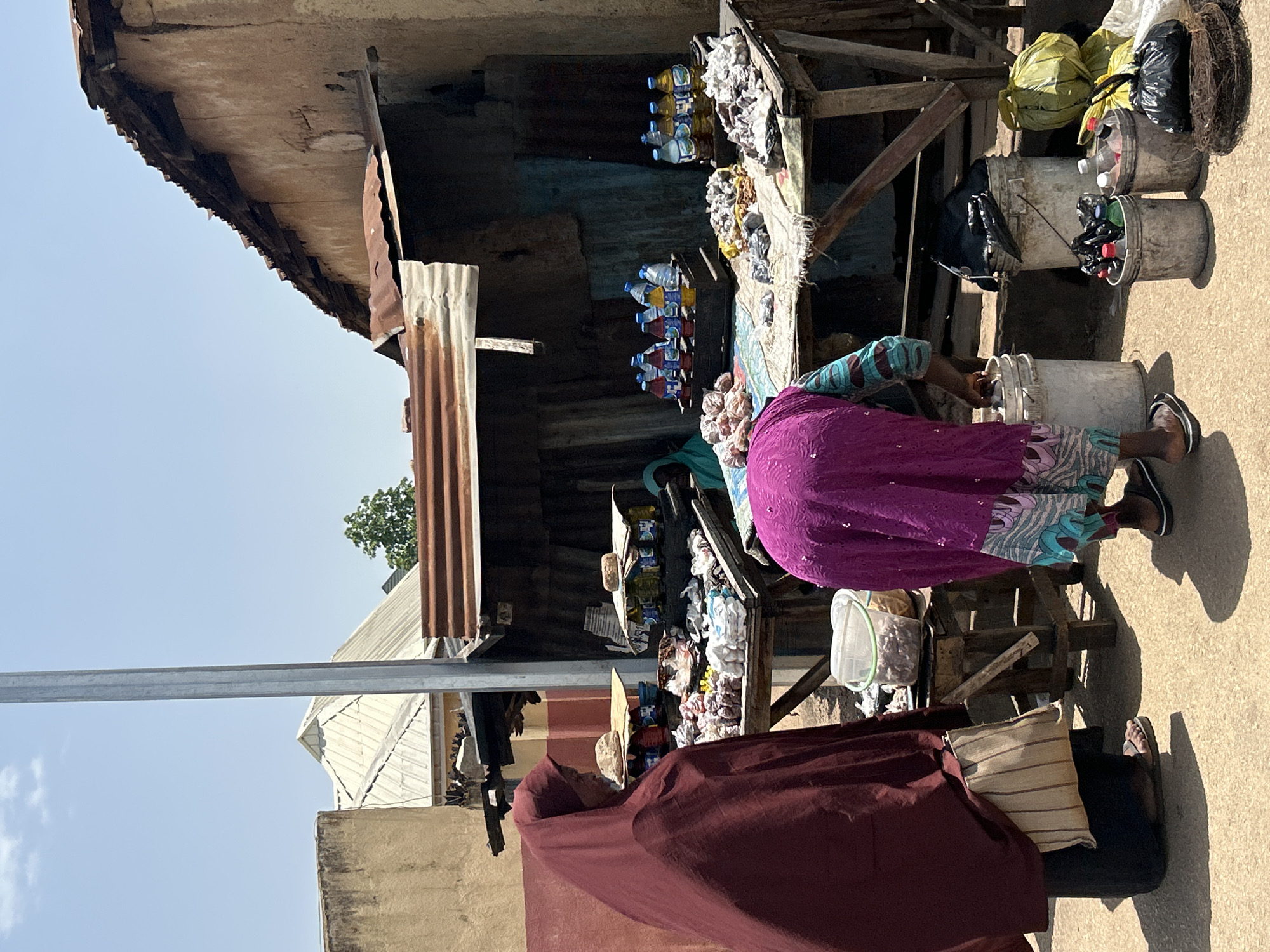 The image depicts an outdoor market scene in Jos, Nigeria, under a clear blue sky. In the foreground, two women are visible. One woman, on the left, wears a dark maroon hijab and a dark skirt, carrying a woven bag. She is standing to the side, facing away from the camera. The second woman, in the center and to the right, is wearing a bright purple abaya with a patterned trim on the sleeves and hem. She is bent over, actively engaged with a white bucket.
The market stall itself is constructed with a corrugated metal roof and wooden planks. It displays various goods for sale, including bottles of drinks (some blue and yellow) arranged in wooden crates on the right side of the stall. There are also piles of what appear to be produce or dry goods on display. In front of the stall, on the ground, are several buckets and bags. The woman in purple is reaching into a white bucket, and next to it are more buckets containing bottles and other items, along with yellow and green plastic bags. A third person, wearing a light green headscarf, is partially visible behind the stall, to the left of the bottles. The ground is paved with a sandy or dusty material, casting shadows from the people and stall. Buildings with earthy-toned walls and corrugated roofs are visible in the background, indicative of the local architecture in Jos. A metal pole, likely part of a street light or utility pole, stands prominently on the left side of the image.