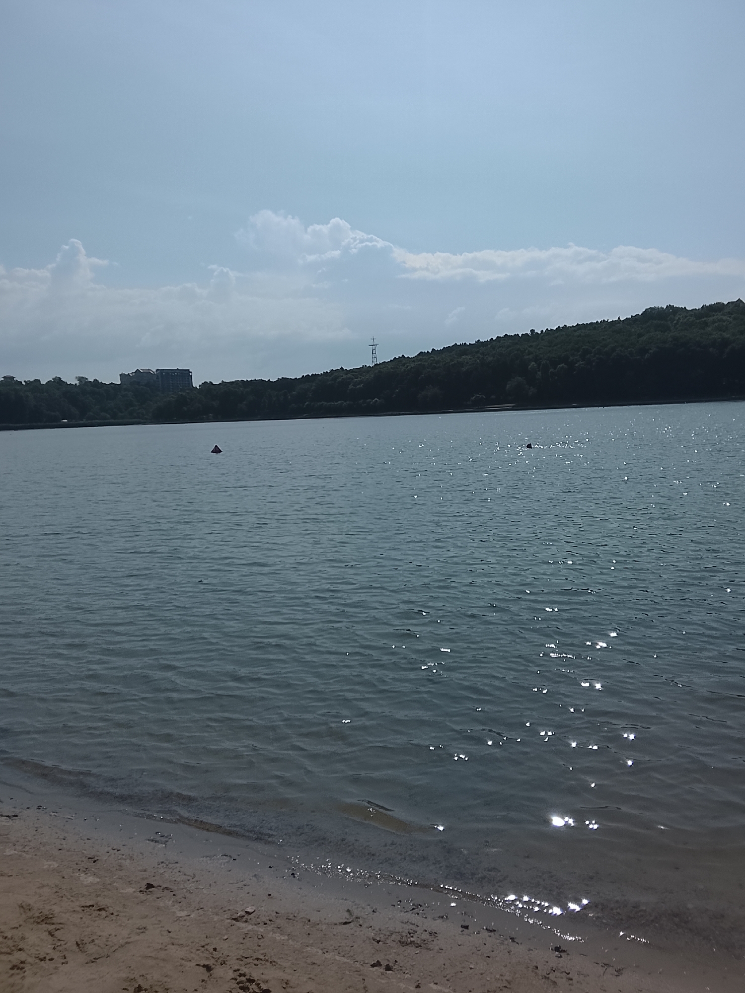 The image depicts a view of a body of water, likely a lake or reservoir, with a sandy shore in the foreground. The water is calm with gentle ripples, and sunlight reflects off its surface, creating shimmering highlights. Across the water, a tree-lined hill forms the background, with a cluster of buildings and a telecommunications tower visible on the ridge. The sky is partly cloudy, with a light blue hue. In the water, there is a red triangular buoy. No people are clearly visible. The scene appears to be a natural or semi-natural landscape within an urban or suburban setting. This location is likely Lake Valea Morilor in Chișinău, Moldova, given its characteristic features of a large body of water, surrounding green hills, and visible urban development on the horizon, as well as the presence of a buoy.