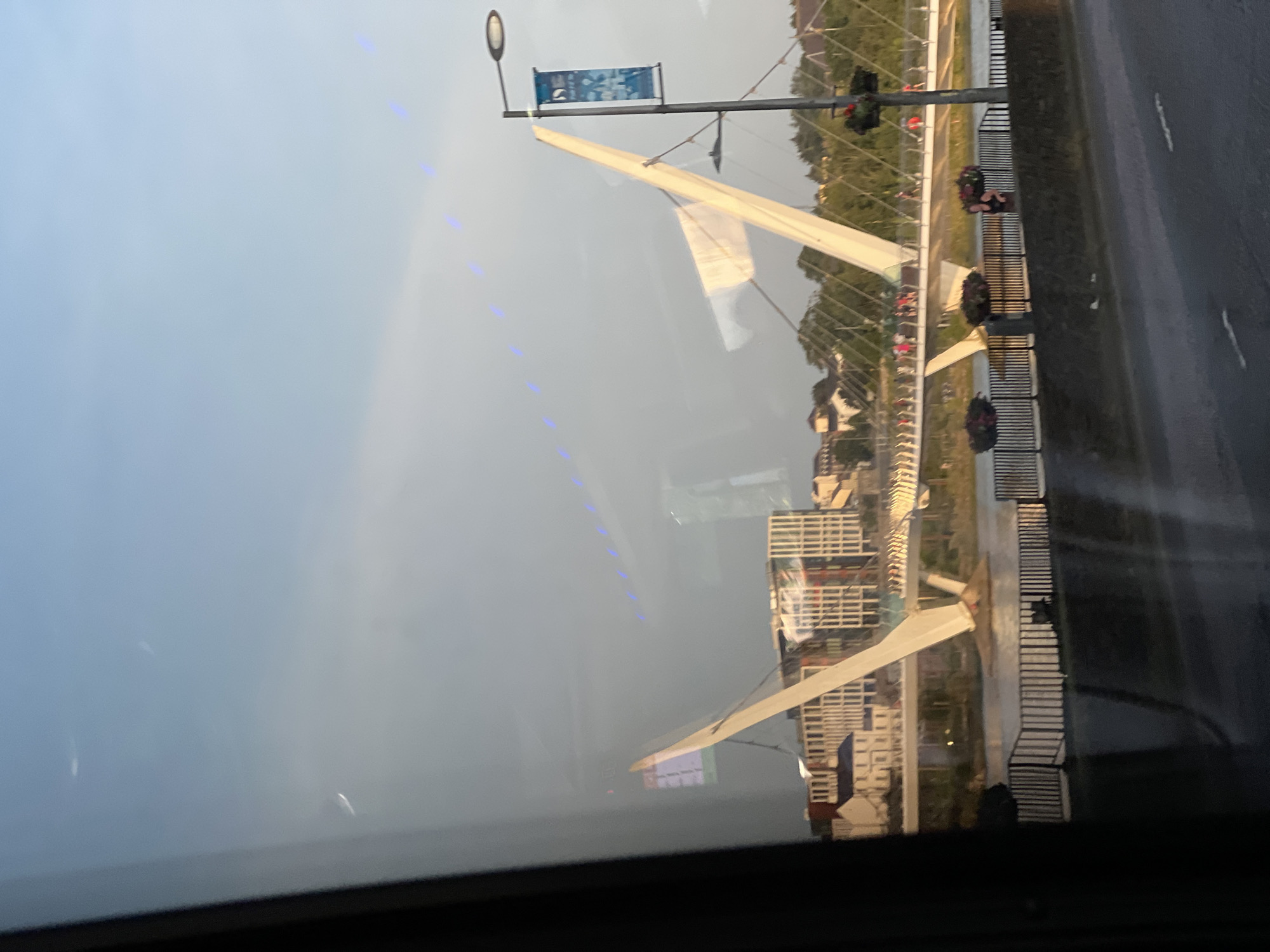 The image captures a reflection of the Peace Bridge in the city of Derry/Londonderry, United Kingdom, viewed through a window. The bridge is a prominent pedestrian and cyclist bridge spanning the River Foyle. The reflection shows the iconic white cable-stayed design of the bridge with its two inclined pylons.
In the background, there are modern buildings, suggesting an urban setting. The sky is partly cloudy, with a soft, diffused light. There are small, blue lights visible in a line across the sky, possibly from a decorative lighting feature or a reflection.
On the bridge itself, several people can be seen walking, indicating it is open and being used. Flower baskets hang from a lamppost on the right side of the image, adding a touch of color to the scene. The foreground shows a dark, reflective surface, consistent with a window, with some smudges or reflections visible. A road surface with lane markings is also visible in the lower part of the image, implying the viewpoint is from a vehicle or near a road. The overall impression is a peaceful, urban scene bathed in gentle light, with the Peace Bridge as the central architectural element.
