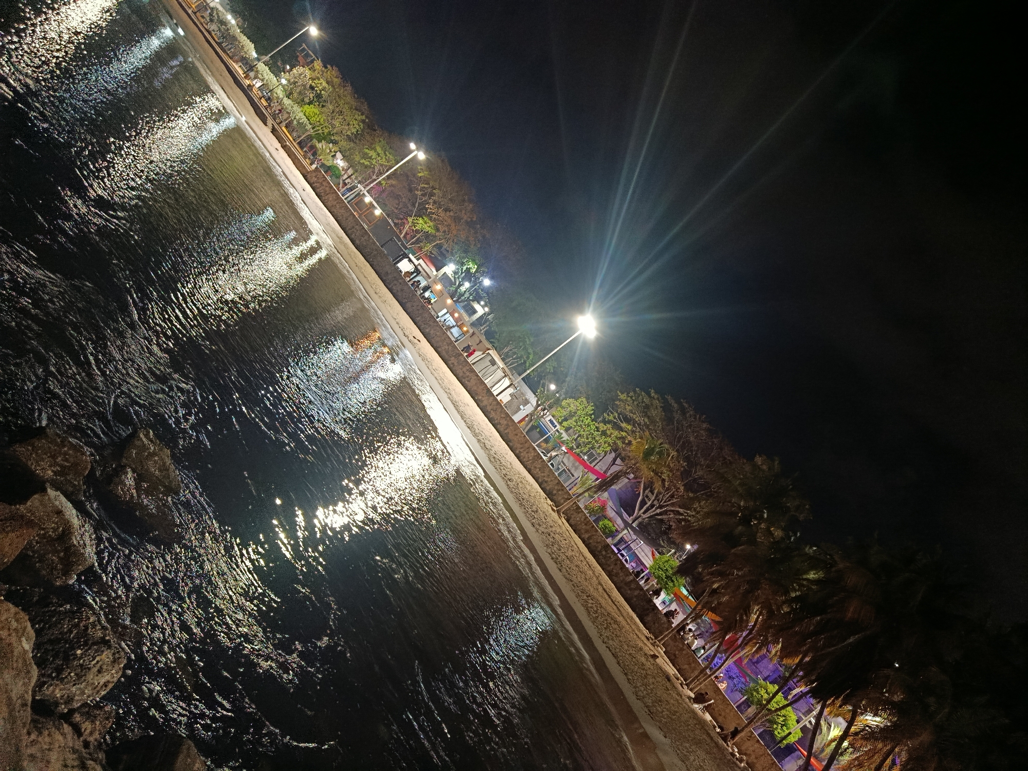 A nighttime view along the coast in Juan Griego, Venezuela, reveals a scene dominated by water and artificial light. A stone embankment separates the water's edge from a sandy strip of beach. Beyond the beach, a walkway lined with trees and streetlights runs parallel to the shoreline. The water reflects the artificial lights, creating a shimmering effect. Several people are visible walking along the walkway. Palm trees are interspersed among the other trees. The sky is dark.