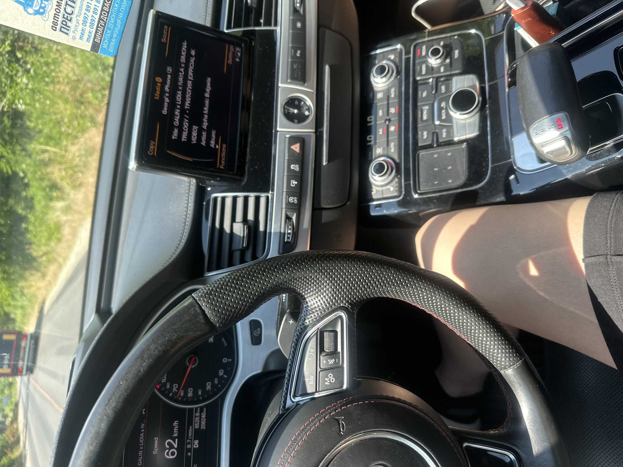 This is a vertical, high-angle shot taken from the driver's seat of a car. The driver's left leg and a portion of their torso are visible in the lower right of the frame, suggesting they are wearing shorts. The focus is on the car's interior dashboard and steering wheel.
The dashboard features a central infotainment screen displaying song information: "GALIN x LIDIA x IVAYLA X SIMONA- TRILOGY / - ТРИЛОГИЯ [OFFICIAL 4K VIDEOJ" by "Alpha Music Bulgaria". The media source is listed as "Georgi's iPhone (2)". Below the screen, there are various car controls, including buttons for hazard lights, traction control, and possibly parking sensors. To the right of the screen is the gear shift, showing "P R N D/S". The steering wheel is black with red stitching and has integrated controls for navigation, volume, and other functions. The speedometer and other gauges are partially visible, with the speed indicated as 62 km/h.
A business card with Bulgarian text is visible at the top left, providing contact information for a business in Ruse, Bulgaria, offering services such as installation, dismantling, and tire services. The scene appears to be set during the day, with bright sunlight casting shadows on the dashboard and the driver's leg. The exterior view through the windshield shows a road and some greenery, with a truck visible in the distance. The overall impression is a snapshot of a car journey in Bulgaria.