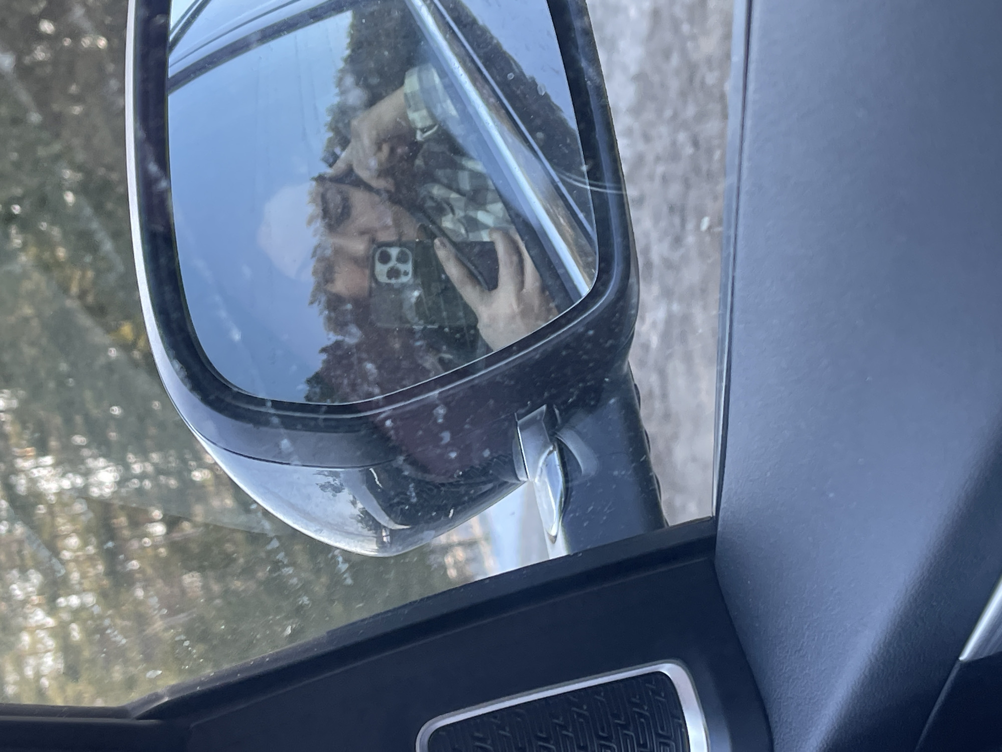 The image displays the exterior side-view mirror of a vehicle, viewed from the interior passenger compartment, located in Kirillovka, Russia. The dark-colored mirror, showing signs of dirt and dust, reflects the upper body of an individual. This person, wearing a green and beige plaid shirt, holds a black-cased smartphone with a distinctive triple-lens camera module in their left hand, directed towards themselves. Their right hand is positioned near their temple. The reflection includes a light blue sky and blurred tree foliage in the background. Through the vehicle's side window, behind the mirror, out-of-focus trees and a general outdoor environment with a gray ground surface are discernible.