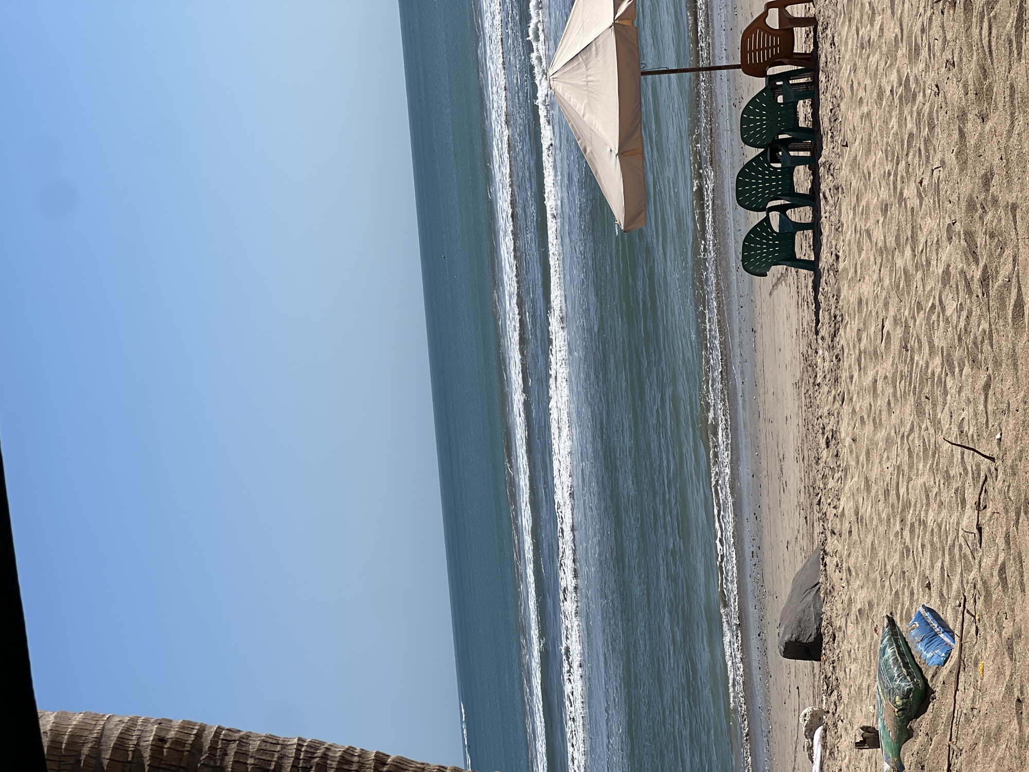 This is a photograph of a beach scene on a clear day. The ocean, with its gentle waves, occupies the upper half of the frame. In the foreground, the sandy beach is littered with debris, including a pillow and a small plastic bag. To the right, an umbrella provides shade for four plastic chairs: three green ones lined up and one brown one to the right of them. The presence of a palm tree trunk on the left edge of the image suggests a tropical location. While the image itself does not explicitly indicate the city of Kuta, Indonesia, such beach scenes are characteristic of coastal areas there.