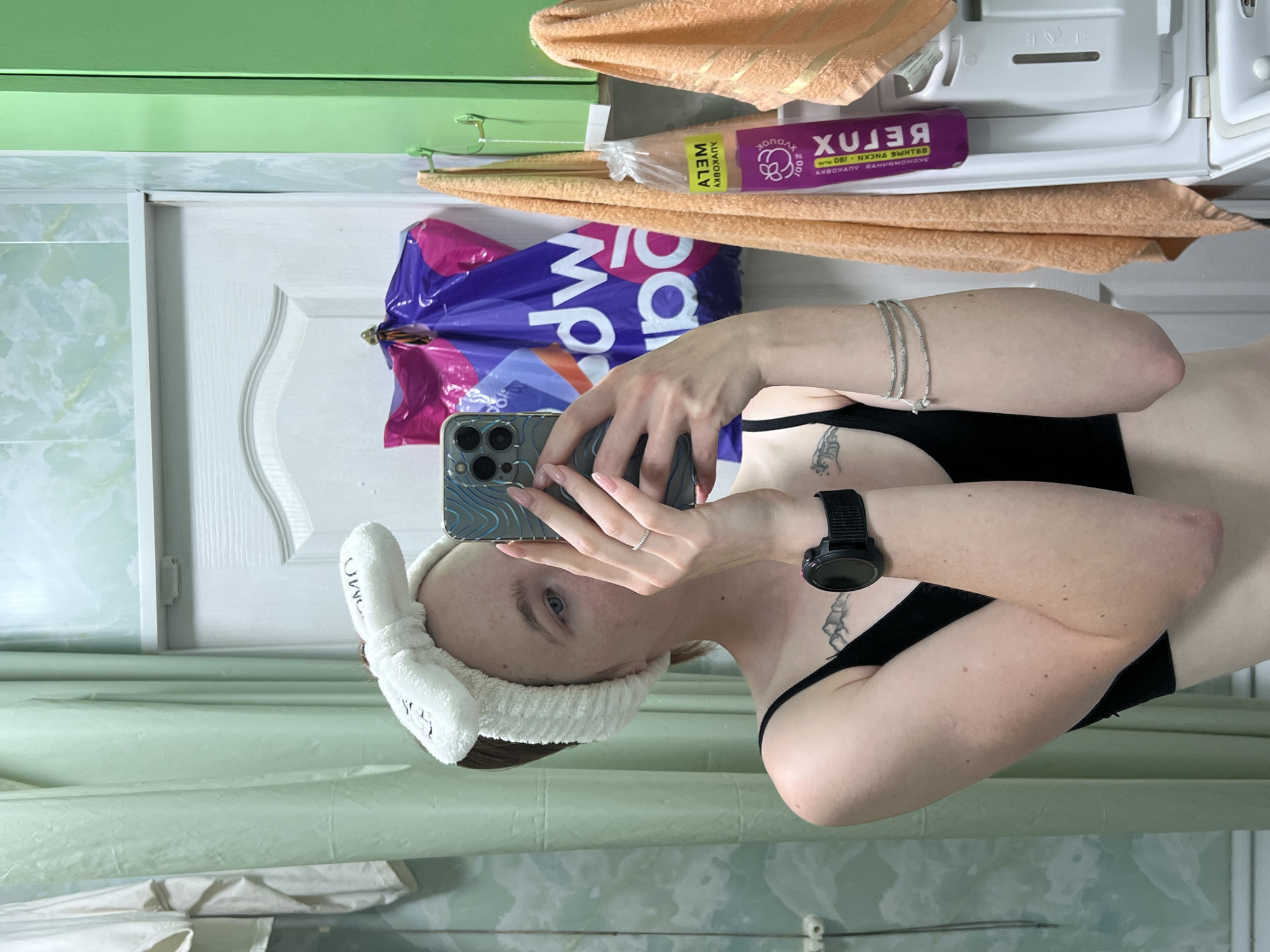 A young woman is taking a mirror selfie in a bathroom, wearing a white fluffy headband and a black sports bra. She has a tattoo on her chest and a watch on her left wrist. In the background, there is a light green shower curtain, a white door, and a green cabinet. A purple shopping bag with white lettering and two orange towels are hanging on the wall. The scene is likely in the city of Chaikovsky, Russia, given the text on the purple product in the background, which appears to be in Cyrillic script.