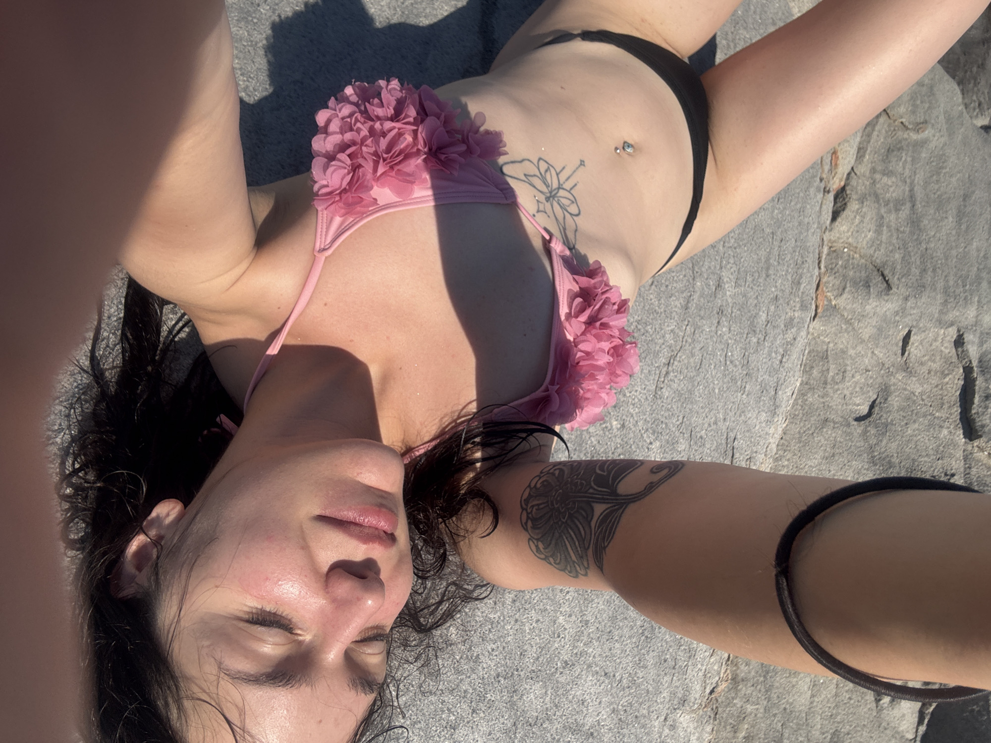 A young woman with dark hair is lying on her back on a rocky surface, possibly near the sea in Chernomorets, Bulgaria. She is wearing a pink floral bikini top and black bikini bottoms. Her eyes are closed, and her face is tilted slightly upwards, illuminated by sunlight. She has a tattoo on her left arm, a butterfly tattoo on her abdomen, and a belly button piercing. A black hair tie is visible on her left forearm. The rocky terrain suggests an outdoor setting, potentially a beach or a coastal area.