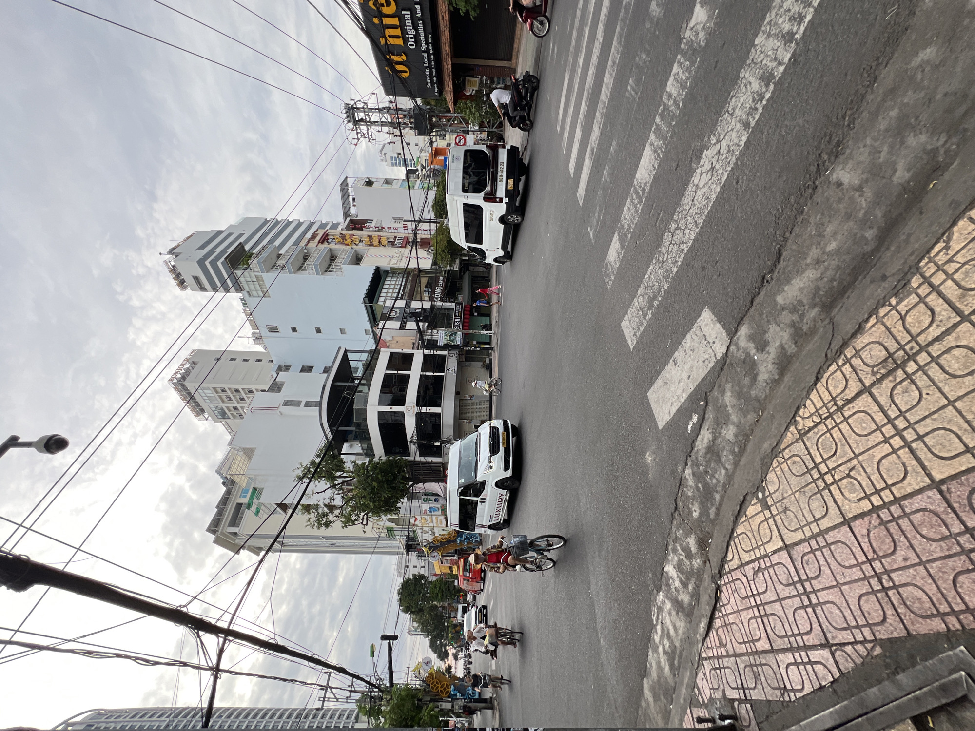 This image captures a bustling street intersection in Nha Trang, Vietnam. The scene is characterized by a mix of modern architecture and urban infrastructure. In the foreground, a pedestrian crossing is marked on the asphalt road, with a patterned tiled sidewalk to the left. Several vehicles are present, including a white van labeled "LUXURY" with license plate "59H-542.23", and another white van. A person on a motorcycle is visible in the middle ground, as is a person riding a bicycle. Multiple buildings with varying architectural styles line the streets, some appearing to be hotels or commercial establishments. Overhead, a dense network of electrical wires crisscrosses the sky, with a traffic light and a surveillance camera visible amongst them. The sky is overcast, suggesting a cloudy day. The overall impression is of a busy urban environment typical of a Vietnamese city.