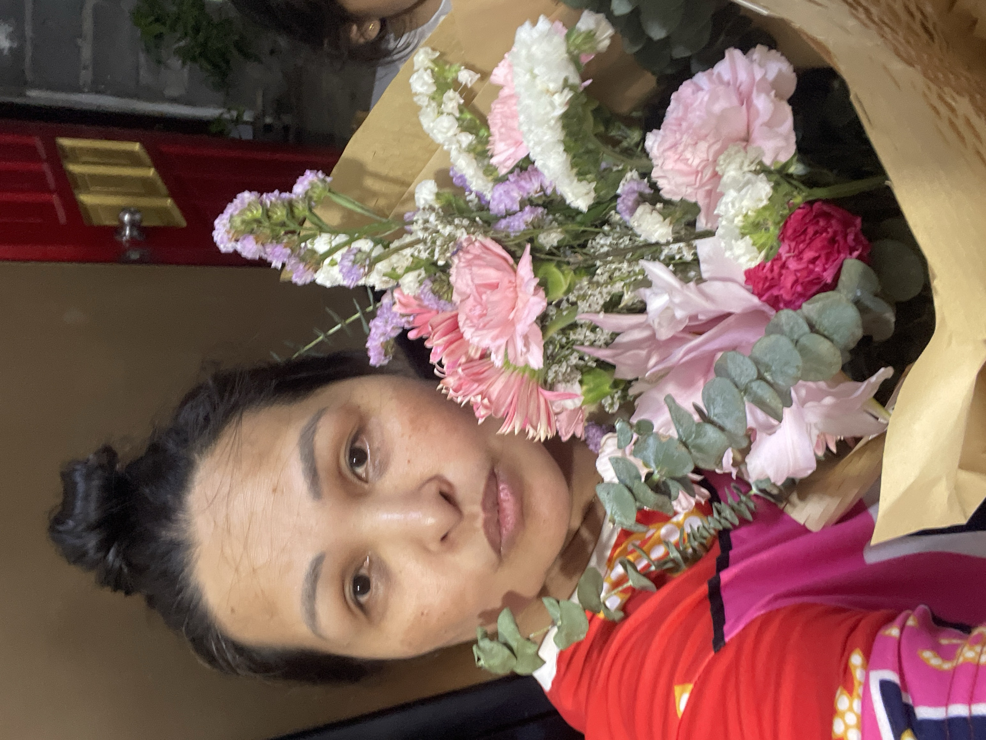 A woman with dark hair pulled back in a bun holds a bouquet of pink, white, and purple flowers wrapped in brown paper. She is wearing a red and pink patterned top. Behind her is a red door with gold accents and a brown wall. The location is Davao City, Philippines.