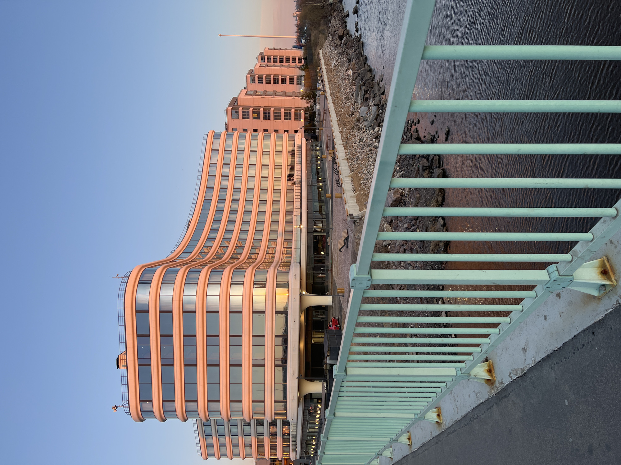 A horizontal medium shot captures a cityscape in Saint Petersburg, Russia, showcasing modern buildings and a river. The scene is bathed in soft, warm light, suggesting either dawn or dusk. The focal point is a contemporary building with a curved façade and reflective windows, adding to the city's architectural diversity. Adjacent to this, more traditional buildings with multiple windows stretch along the riverfront. A metallic bridge railing in a light green color runs alongside the view, hinting at the vantage point of the photo. The river, which is close to the building, appears dark and still, reflecting the light from the sky and buildings. The sky transitions from a light blue to a soft pink hue, indicating the time of day. The composition of the shot, with the railing in the foreground and the cityscape in the background, creates depth and perspective.