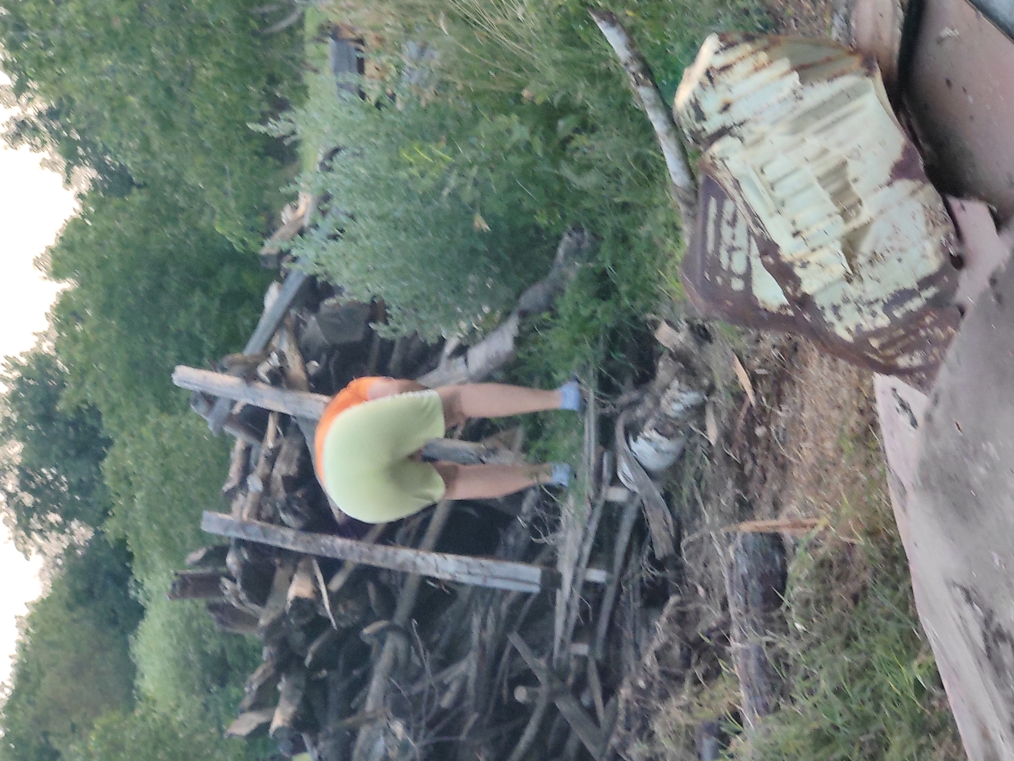 The image depicts a person, seen from behind, wearing light green shorts and an orange shirt, bending over near a pile of wood. The person is wearing blue shoes. The background consists of trees and dense green foliage. In the foreground, there is a large piece of rusted metal. The scene appears to be outdoors, possibly in a rural or overgrown area. The location is stated as Kochmar, Bulgaria.