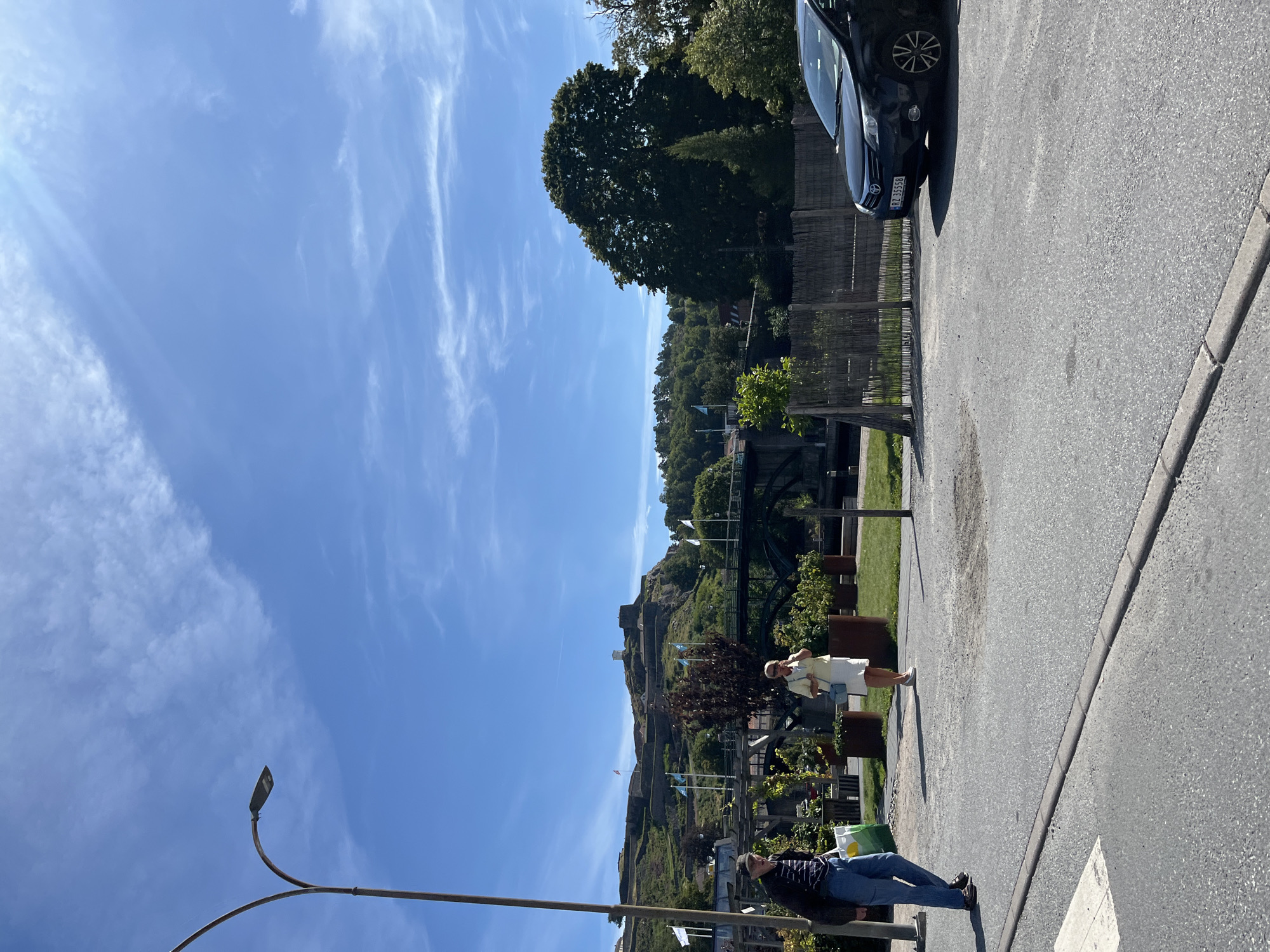 The image depicts a street scene in Halden, Norway, with the imposing Fredriksten Fortress visible on a hill in the background. In the foreground, two individuals are walking on a paved area. A man on the left, wearing a striped shirt and jeans, carries a green and yellow shopping bag. A woman on the right, dressed in a light-colored outfit, holds a light blue purse. A dark-colored car is parked on the right side of the road, with its license plate visible as "RZ 35558". The environment features a street, a grassy area with planters, a wooden fence, trees, a bridge over a body of water, and flags displayed on poles. The sky is blue with scattered white clouds. The lighting suggests it is daytime.
