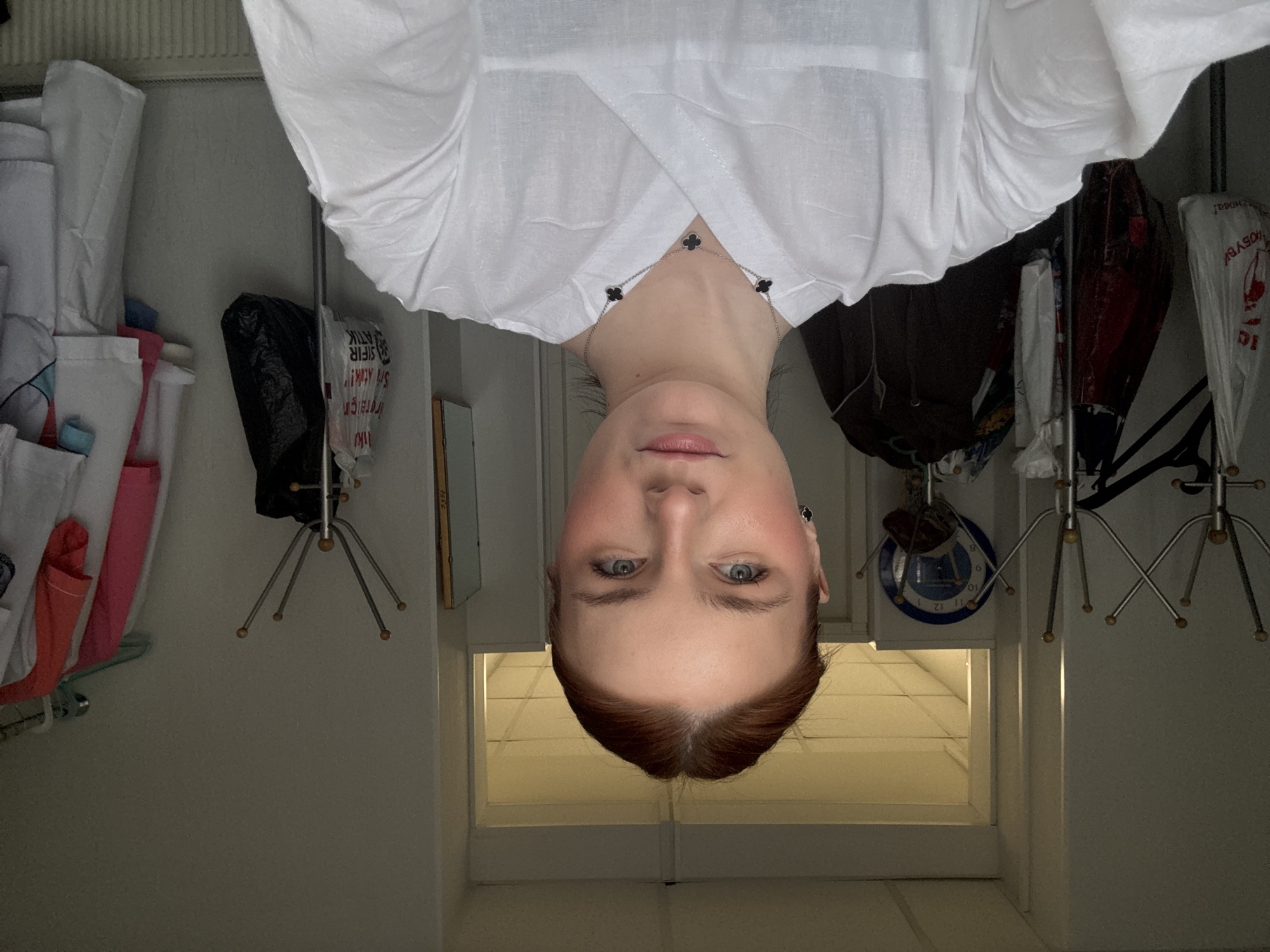 The image presents an inverted view of a fair-skinned person with red hair wearing a white top. A clover-shaped necklace rests just below their chin. The background includes various items hanging on racks or hooks, such as bags, clothing, and cleaning supplies. In the background, there is also a white door and a wall clock. The location of the photo is not provided in the image. The image is not specifically connected to St. Petersburg, Russia.