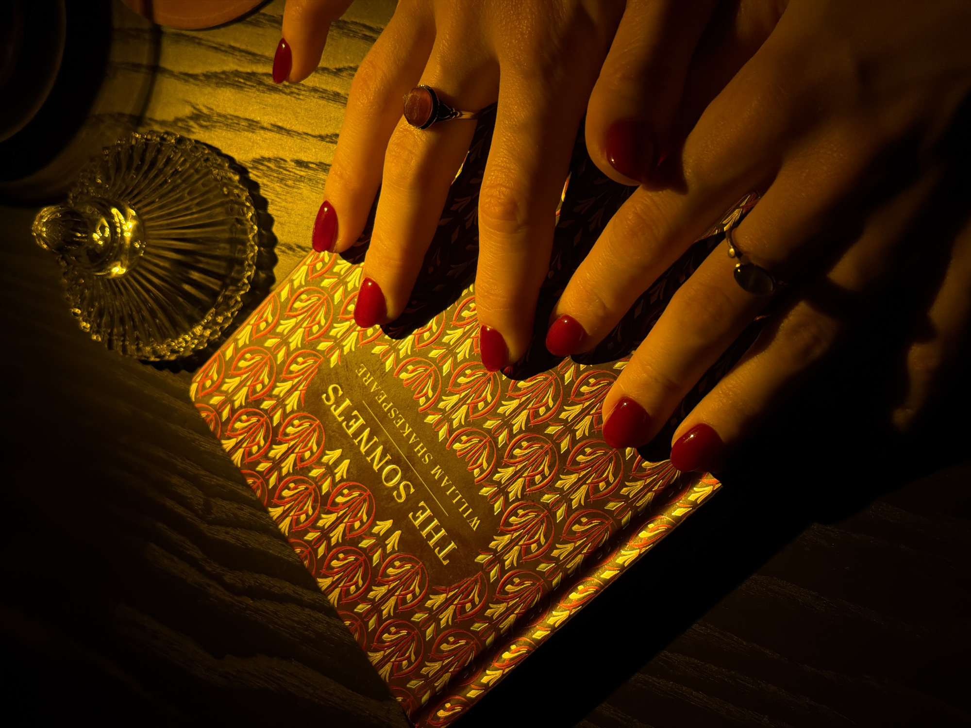 The image depicts two hands with red manicured nails resting on a book titled "The Sonnets" by William Shakespeare. One hand has a ring with a red stone on the middle finger, and the other has a ring on the pinky finger. To the left of the book is a small, decorative glass dish. The scene is dimly lit, casting shadows on the wooden surface. The location in the city of Đà Nẵng, Vietnam is not discernible from this image.