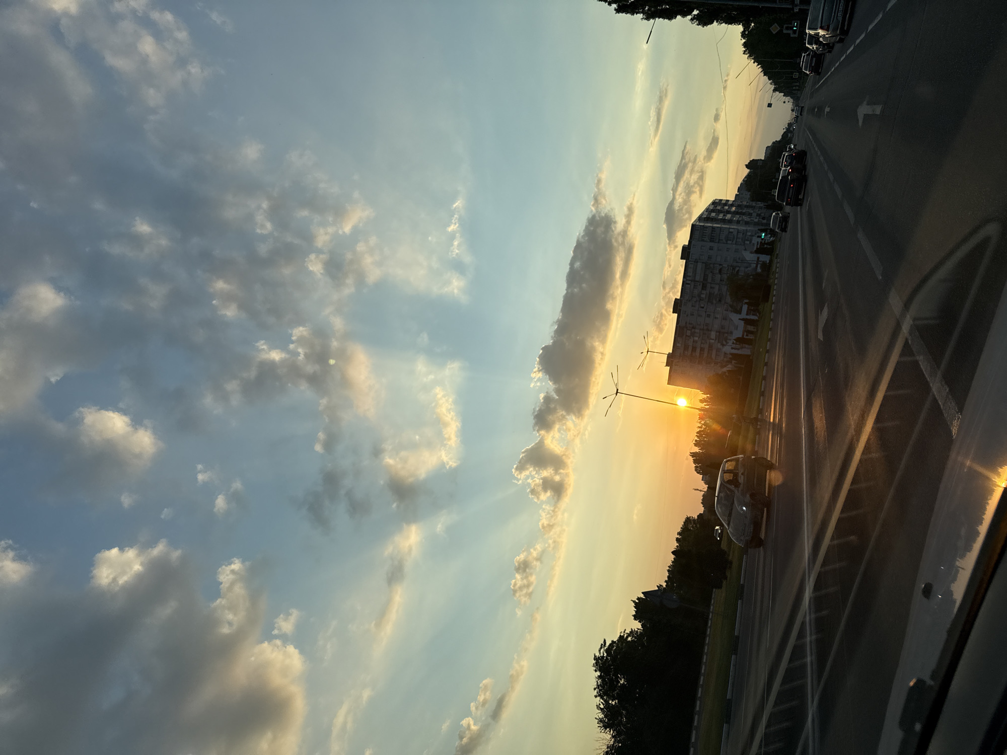 The image depicts a view from a vehicle, likely in Stary Oskol, Russia, during sunset. The scene is dominated by a dramatic sky filled with clouds, some of which are illuminated by the setting sun, creating a warm, golden glow. Rays of sunlight pierce through the clouds, casting a dramatic effect.
On the right side of the image, a multi-lane road is visible with several cars driving on it. The road appears to be a major thoroughfare. In the background, to the left of the road, there is a large, multi-story residential building. Trees line the side of the road and the area around the building.
The time of day is clearly sunset, indicated by the low angle of the sun and the warm, orange and yellow hues in the sky. The weather appears partly cloudy, with a mix of dark and light clouds. The overall mood is serene and picturesque, capturing the beauty of a sunset over an urban landscape. There is no visible text in the image.