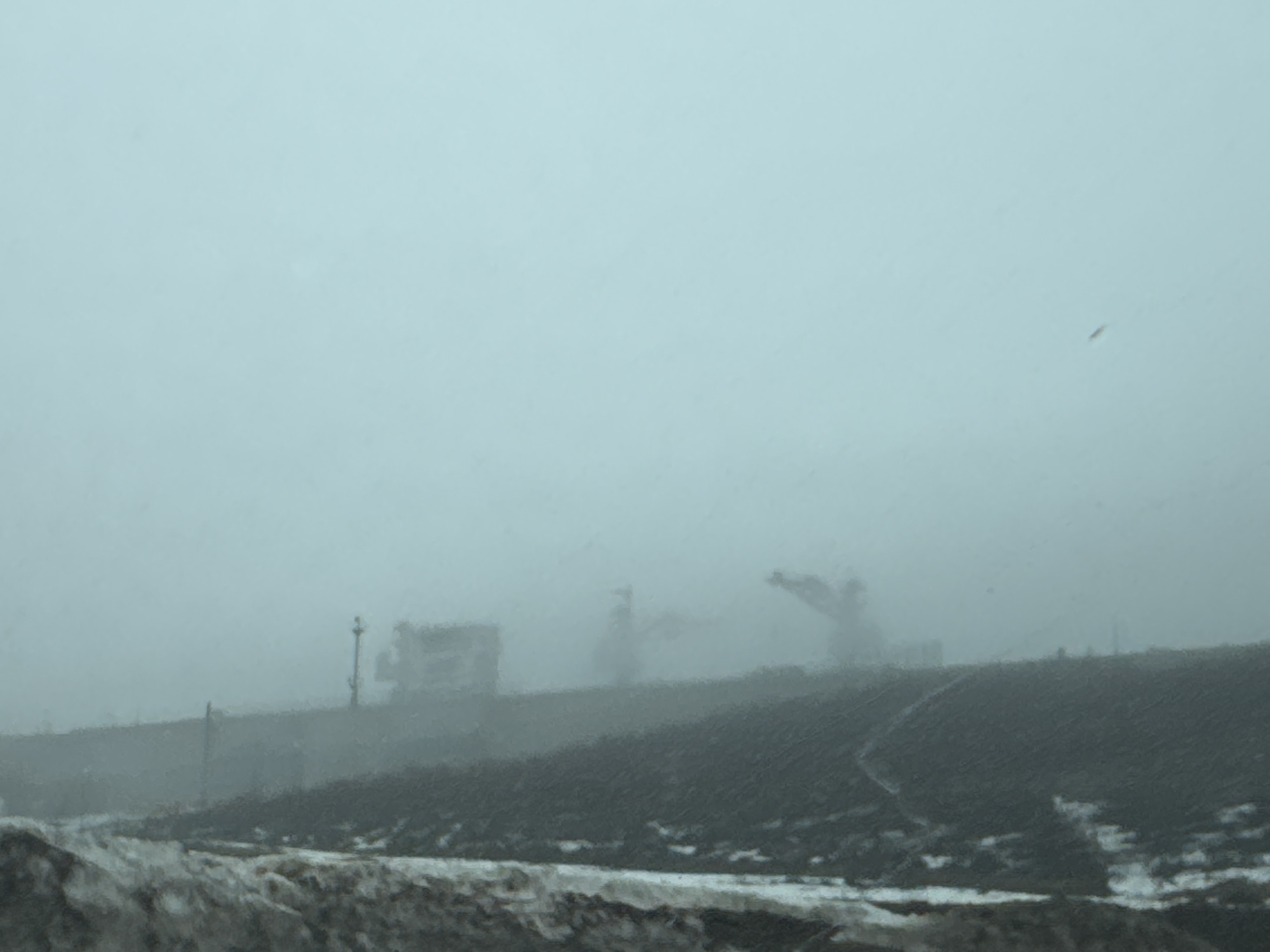 The image depicts a hazy scene in what appears to be an industrial or construction area. Visible are the blurred outlines of heavy machinery, including what looks like cranes and large vehicles. There is a dark, possibly dirt or snow-covered embankment in the middle ground. The background is obscured by fog or snow. Based on the prompt, this scene is located in the city of Chaikovsky, Russia.