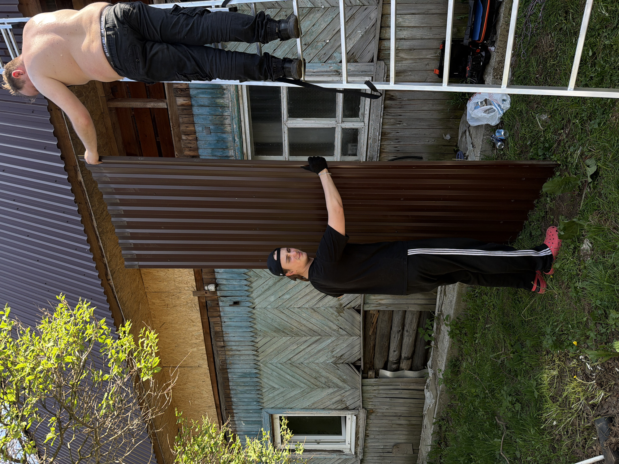 Two men are working on the roof of a dilapidated wooden building. One man, shirtless, is on a white ladder, while the other, wearing a black shirt, pants, gloves, and a cap, is holding a brown corrugated metal sheet in place. The building has a visible window and weathered, peeling paint. The ground around the building has grass and some debris. The location appears to be a rural or less developed area. No specific evidence suggests the city of Perm, Russia.