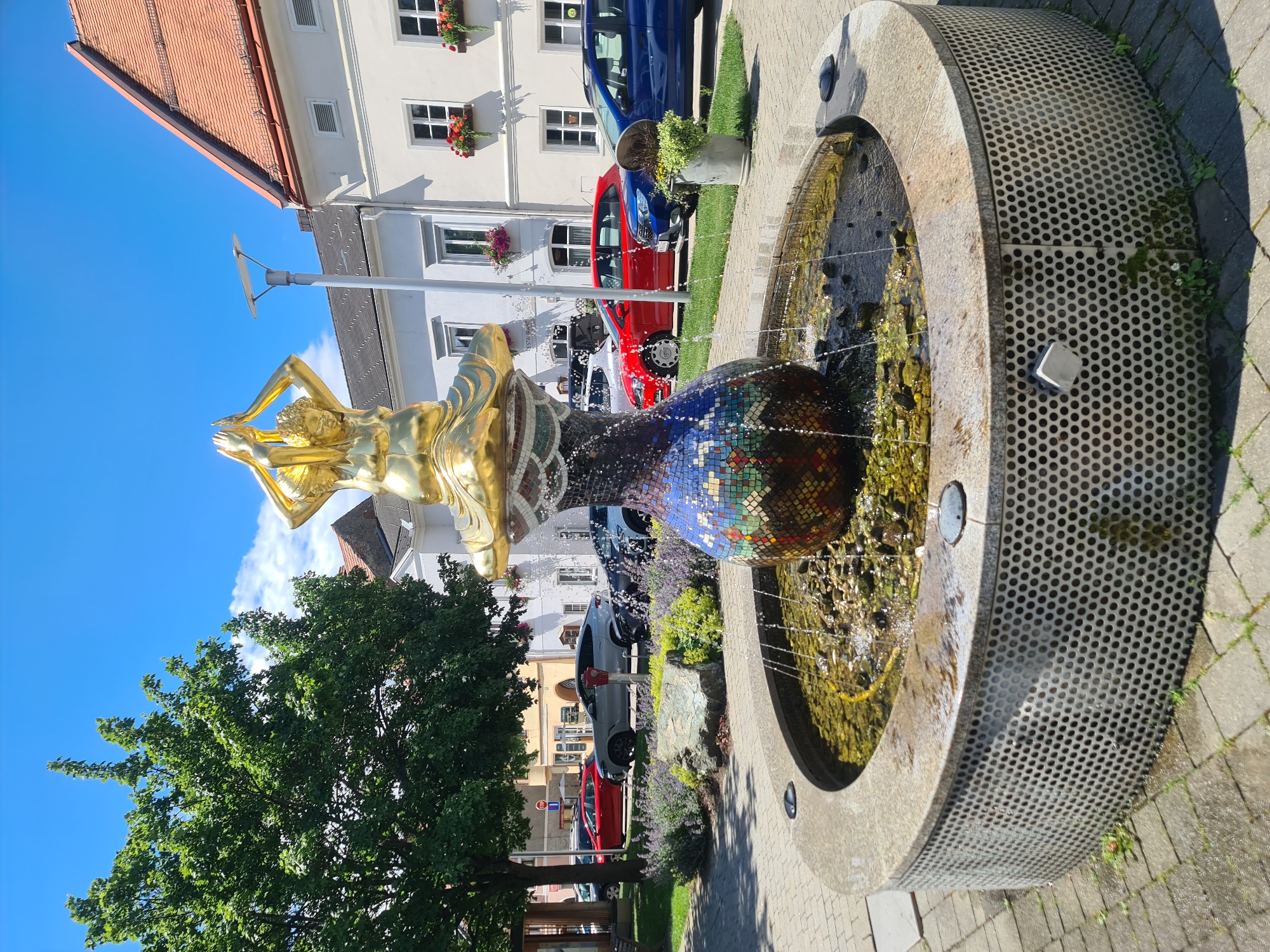 The image depicts a public fountain in Sankt Marein bei Neumarkt, Austria. The fountain features a golden statue of a figure with hands clasped overhead, atop a mosaic-covered spherical base. Water cascades from the statue into the basin, which is filled with water and small stones. The fountain is surrounded by a circular concrete structure with a perforated metal exterior. Paved walkways and greenery, including lavender bushes and a large tree, are visible around the fountain. In the background, buildings with tiled roofs and parked cars line a street. The sky is clear and blue with a few white clouds.
