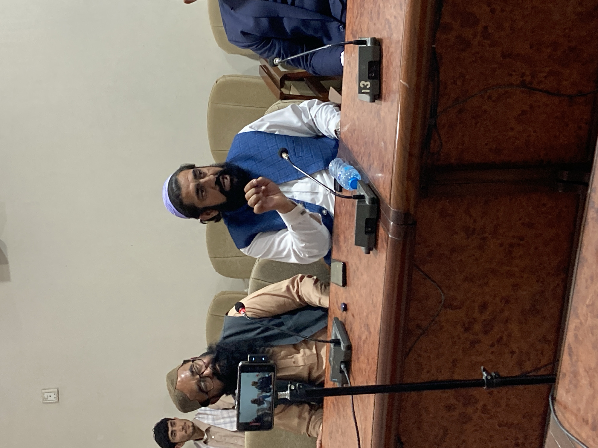 The image depicts a meeting or conference taking place, likely in an official or professional setting. Three men are visible seated at a large wooden table equipped with microphones. The man at the top of the frame is wearing a white shirt and a blue vest, gesturing with his right hand while speaking. Next to him on the table, there is a water bottle and a microphone. Another man below is wearing a tan shirt and a dark vest and has a long beard. His eyeglasses are perched on his nose. A smartphone is mounted on a stand, recording or streaming the event. A third man in a light-colored shirt is partially visible in the foreground. The setting appears to be indoors, possibly in a conference room or hall, with light-colored walls and upholstered chairs. There is no indication of the specific location within Quetta City Tehsil, Pakistan, beyond the general professional/official atmosphere.