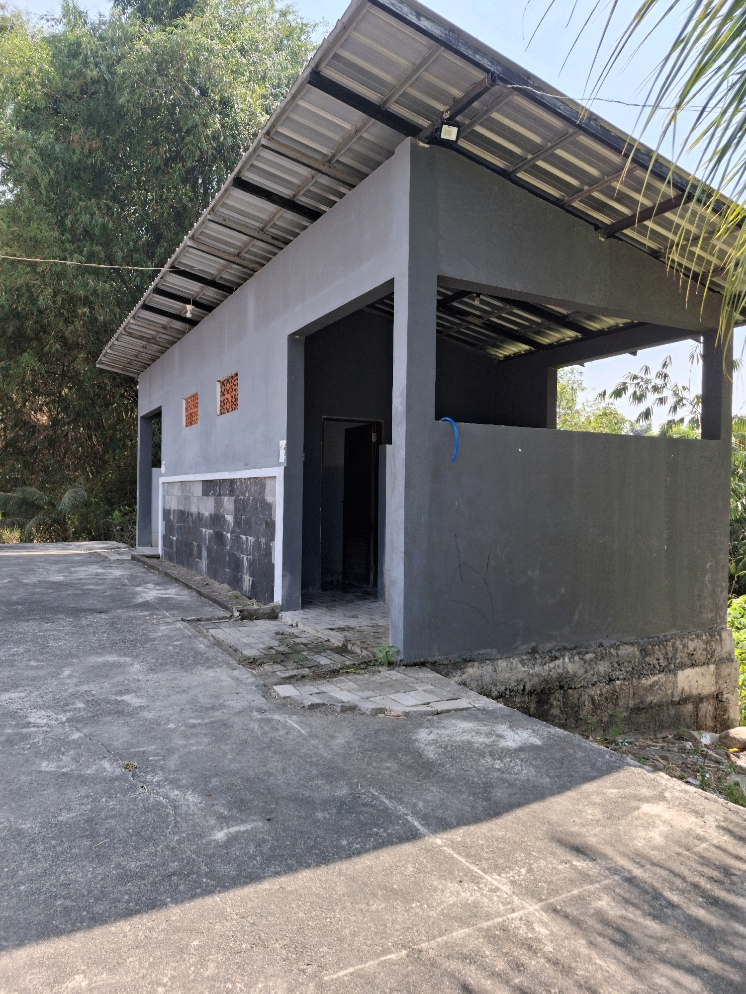 An outdoor, concrete-floored building with a corrugated metal roof, painted gray, is located in Kedungmaron, Indonesia. The building has a rectangular footprint with an open-air section. On the left side of the building, there are two small square windows with orange brick detailing. Below these windows, the lower portion of the wall is covered in dark gray rectangular tiles. A dark-colored door is visible on the right side of the building, leading into an enclosed space. A blue hose is attached to the exterior wall in the open-air section. The ground around the building is paved with concrete, and the area is surrounded by vegetation, including trees and palm fronds. The sky is clear and blue. The image appears to be a photograph taken during daylight hours.