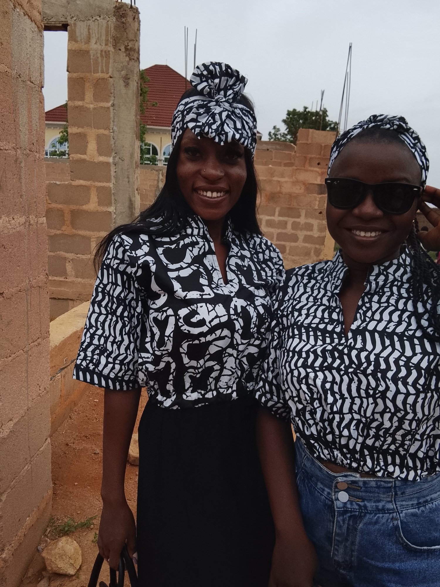 Two dark-skinned women stand shoulder-to-shoulder outdoors. The woman on the left wears a black and white patterned short-sleeved shirt, a black skirt, and a matching black and white patterned head wrap. The woman on the right wears a similar patterned shirt and blue jeans, with a matching head wrap and sunglasses. Both women are smiling. The background consists of an unfinished brick building and a house with a red roof. The location is Barikin Sabon Gidan Kanat, Nigeria.