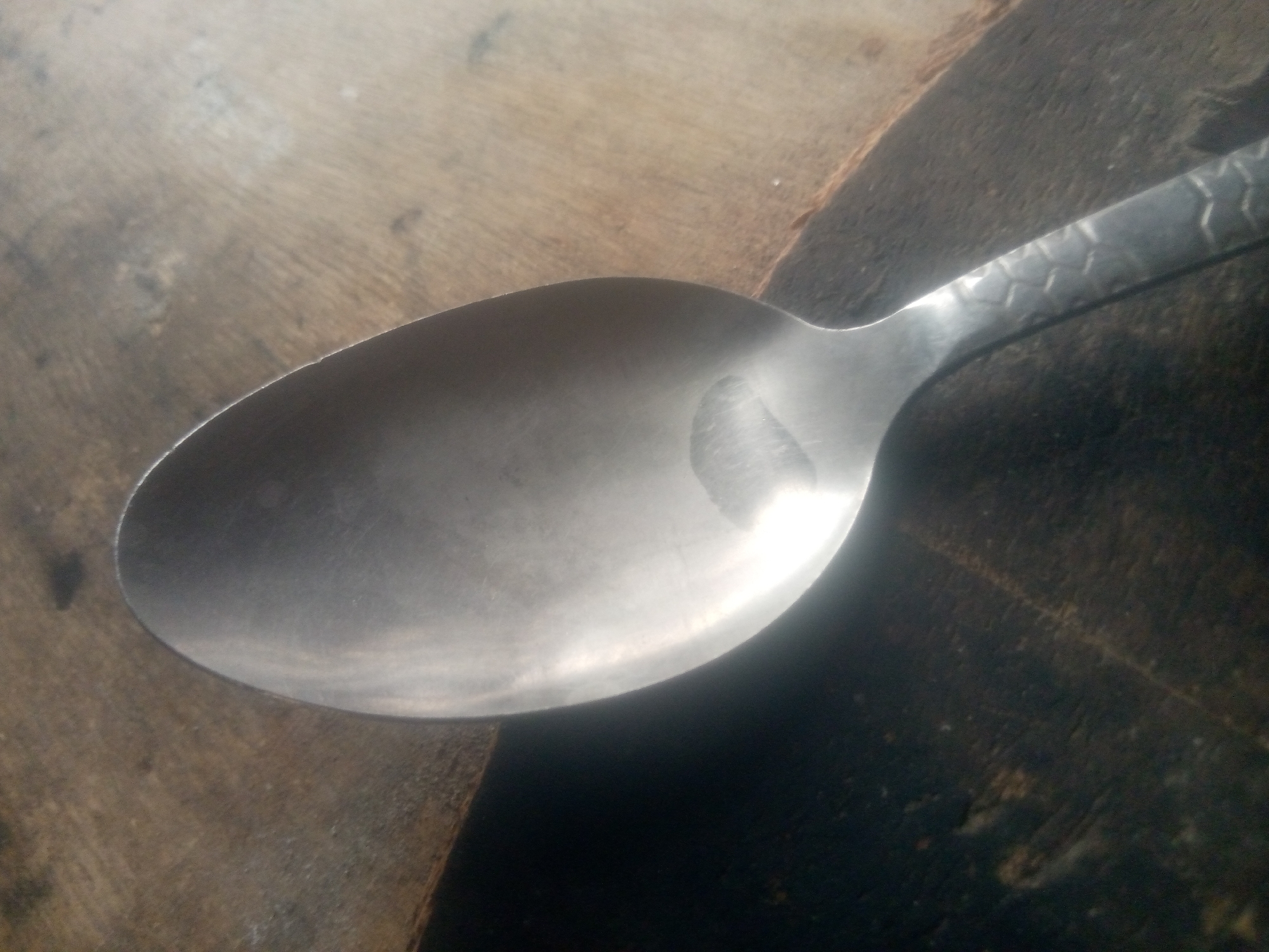 The image shows a close-up of a metal spoon resting on a textured wooden surface. The spoon is angled, with the bowl facing the viewer and the handle extending towards the top right of the frame. The spoon's surface is somewhat reflective, showing a smudged area on the bowl. The handle of the spoon has a textured pattern, resembling small diamonds or scales.
The wooden surface beneath the spoon appears to be a piece of lumber with a visible grain. The background is a dark, out-of-focus area that contrasts with the lighter wood and the metallic sheen of the spoon. The lighting suggests it could be daytime, with some light reflecting off the spoon's surface.
There is no visible text in the image, nor are there any people or animals. The scene is simple and focuses on the object and its immediate surroundings. The context of Jauro Yinu, Nigeria, does not provide specific visual cues within the image itself, but it establishes the geographic location.