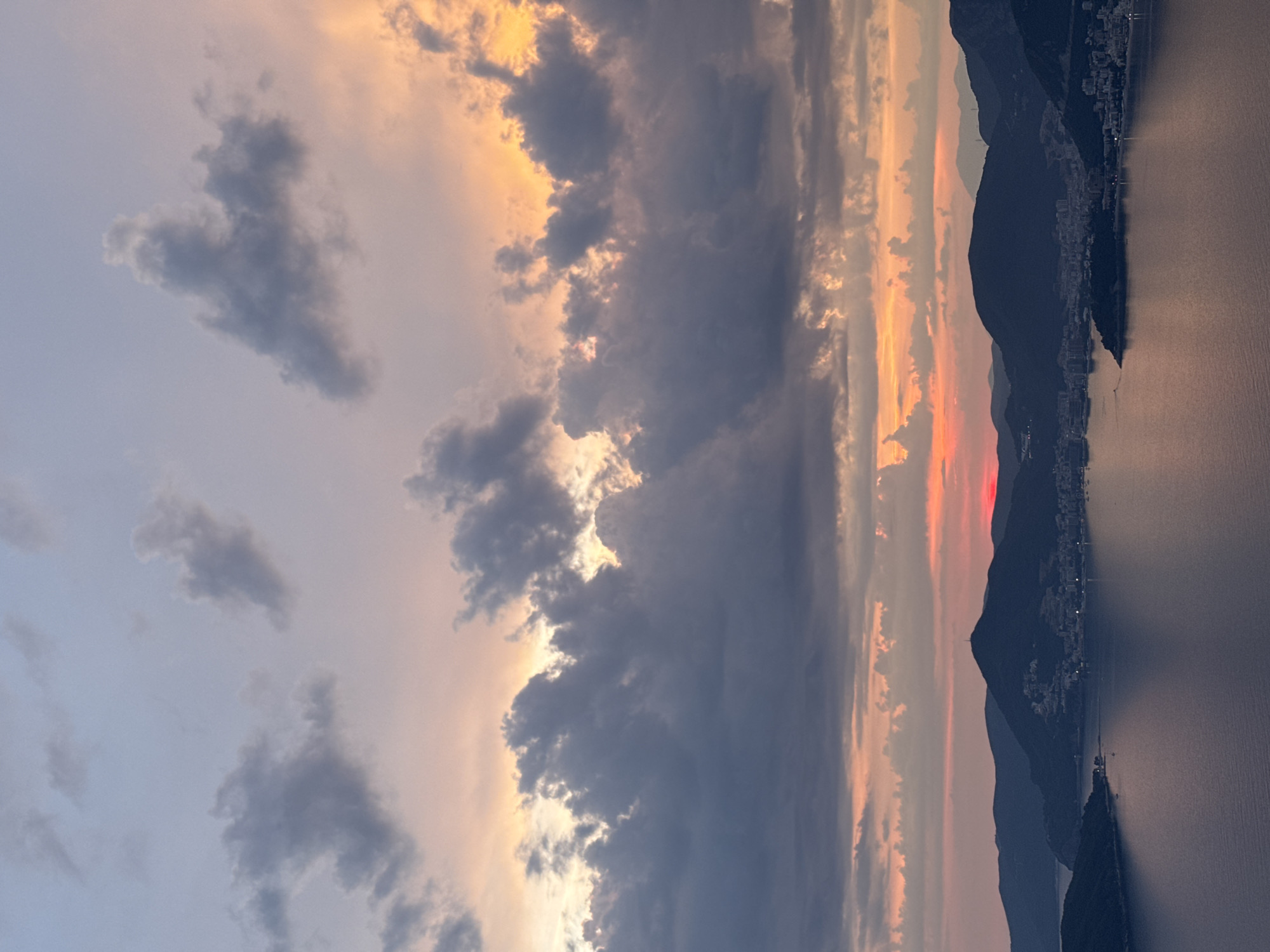 A wide shot captures a twilight scene over the coast of Sveti Stefan, Montenegro. The sky is a canvas of soft pastels, transitioning from a pale blue at the top to hues of pink, orange, and yellow closer to the horizon. Dark, voluminous clouds drift across the sky, their undersides tinged with the warm glow of the setting sun. The sun itself is a tiny, bright red disc, partially obscured by clouds, sinking towards the horizon.
Below the sky, a dark, undulating mountain range silhouettes the coastline. Nestled within the curves of the mountains is the town of Sveti Stefan, a cluster of buildings appearing as small lights against the darkening landscape. The calm water of the sea reflects the muted colors of the sky and the dark forms of the mountains. Several small boats are visible dotted on the water, their shapes indistinct in the low light. The overall impression is one of natural beauty and tranquility, characteristic of a coastal sunset in the region.