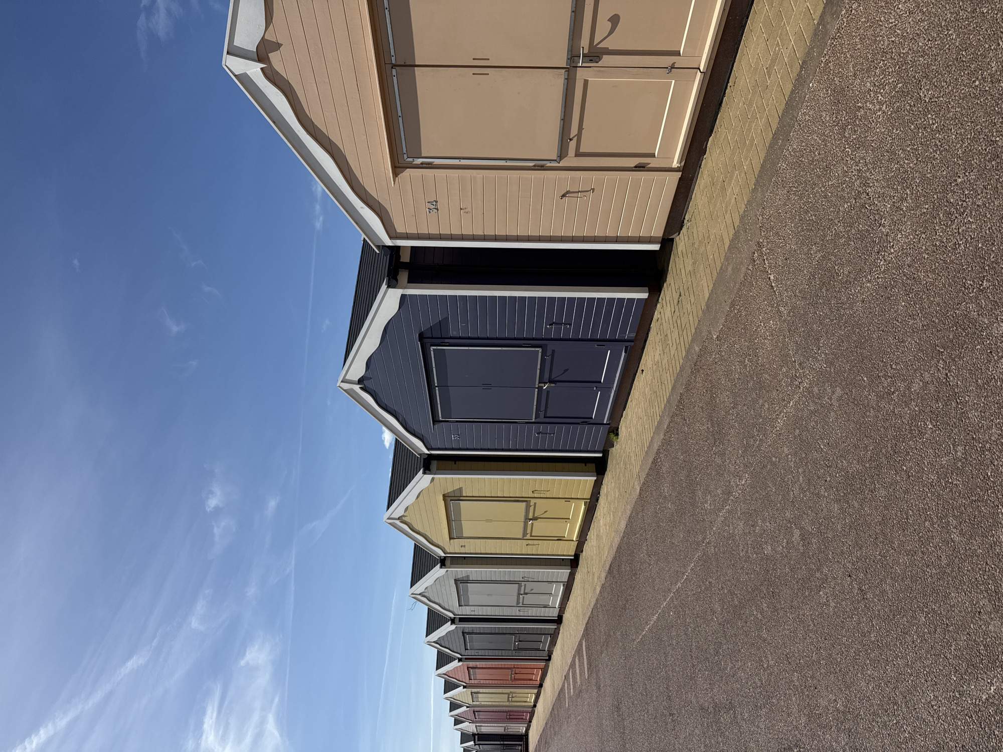 The image shows a row of colorful beach huts along a path, typical of seaside locations. The sky is a clear blue with wispy clouds. The huts are painted in various pastel colors, from peach to grey, and are built in a traditional style. The path next to the huts is paved. The location is Great Yarmouth, United Kingdom.
