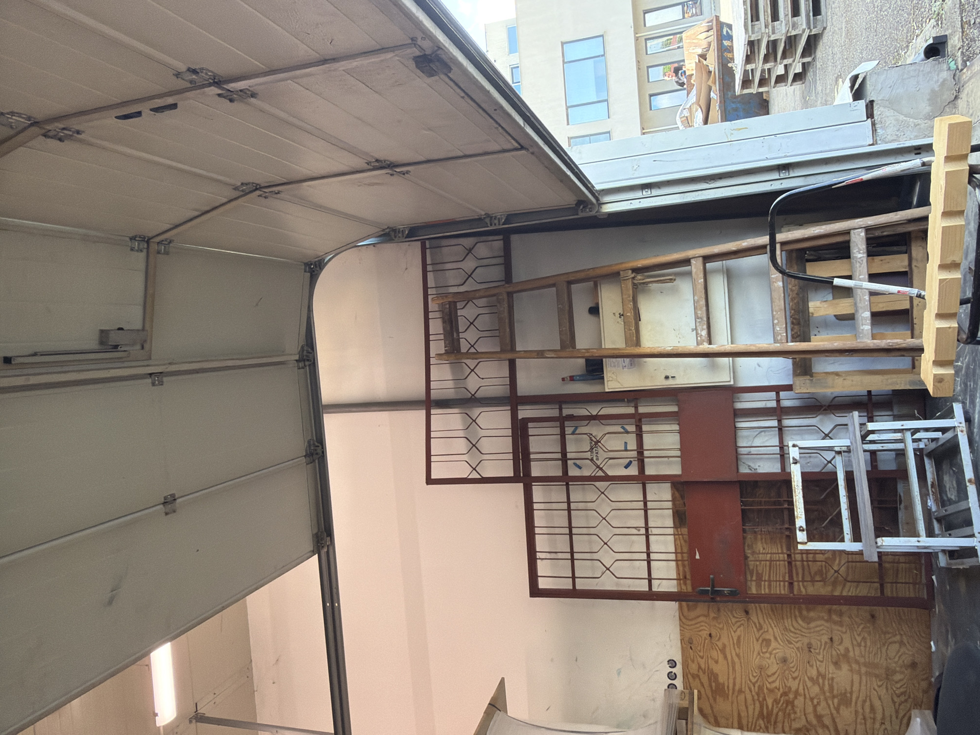 The image shows an interior view of a garage in Ravno Pole, Bulgaria, with the overhead garage door partially open, revealing the exterior. Inside, the garage is cluttered with various items. To the left, a large piece of plywood leans against the wall. In the center, two large, red, metal security gates stand upright, one slightly overlapping the other. In front of these gates is a small, white metal shelf unit on wheels. A wooden ladder is positioned to the right of the gates, leaning against the wall. Further to the right, a metal cabinet or electrical panel is mounted on the wall. In the foreground, a piece of wood with notches is placed on a dark surface. Outside the garage, several wooden pallets are stacked, and a blue dumpster is visible. A person wearing a yellow hard hat is seen in the background near the dumpster. The overall environment appears to be a workshop or storage area.